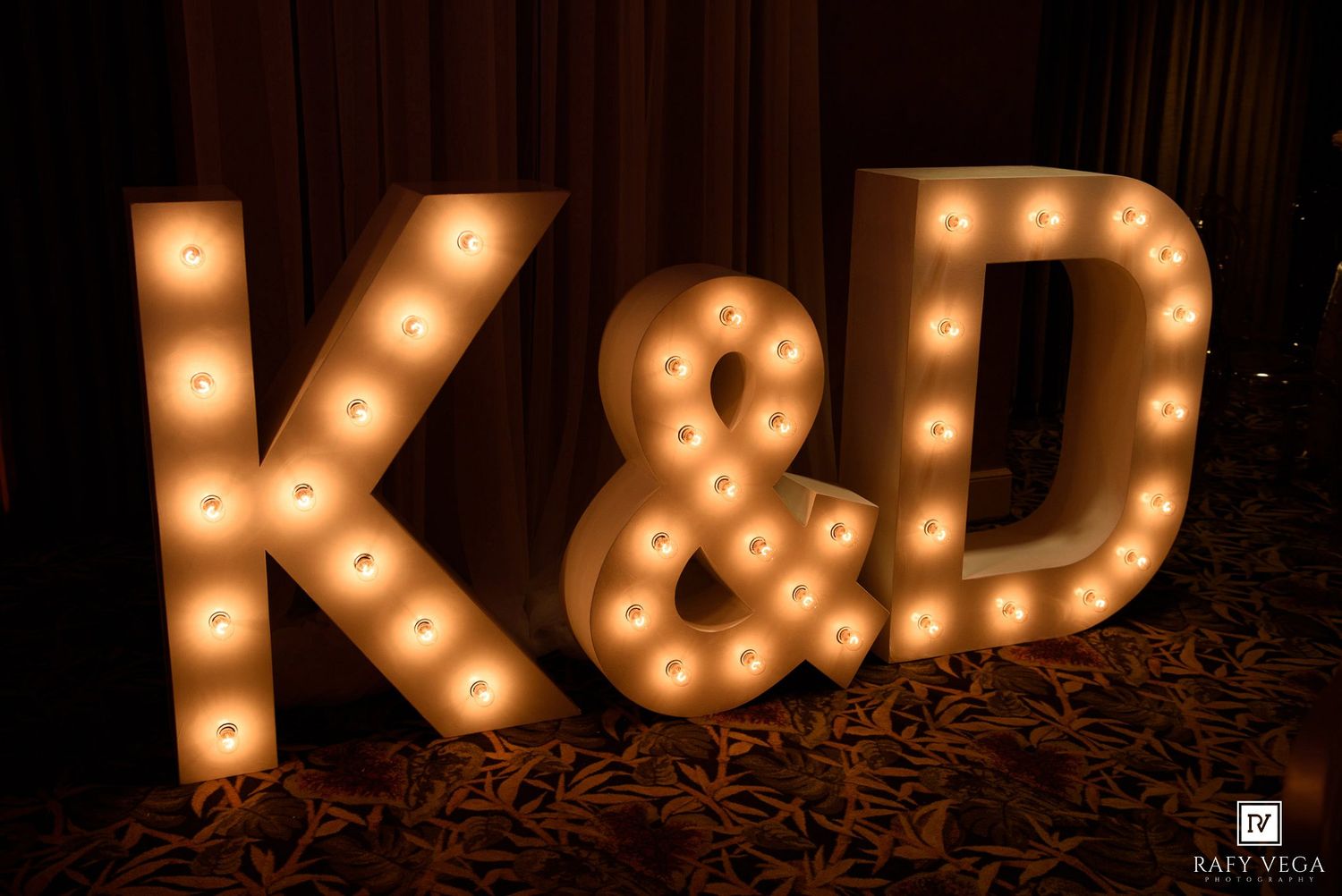 Illuminated marquee letters K&D light up a wedding reception venue with warm bulb lighting.
