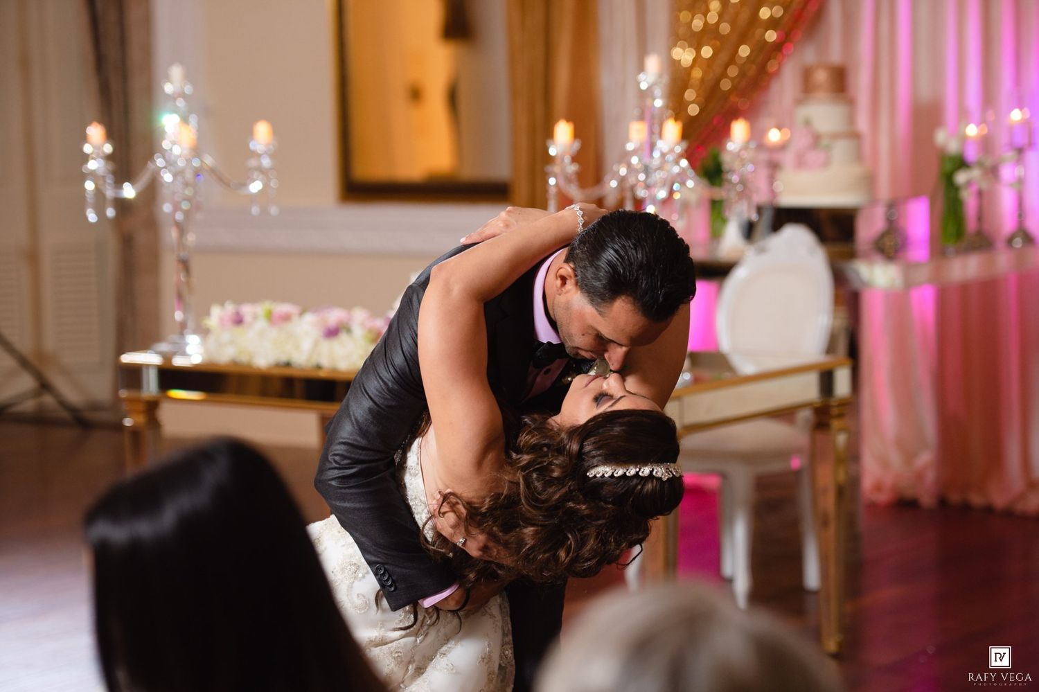 Bride and groom share a romantic dip kiss at their elegant wedding reception with pink lighting and chandelier decor.