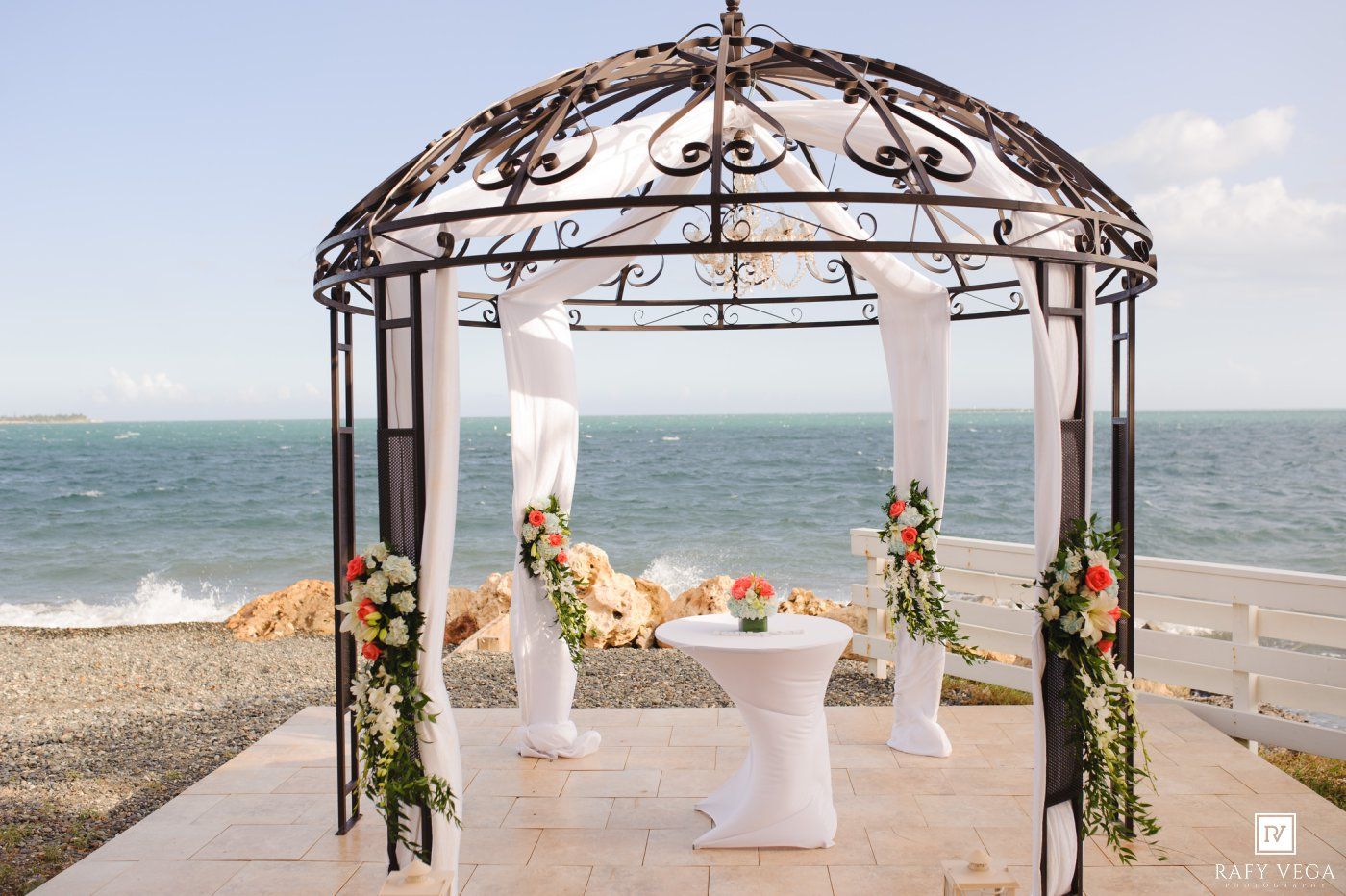 Elegant iron gazebo wedding ceremony setup with white drapes and floral arrangements overlooking a scenic ocean backdrop.