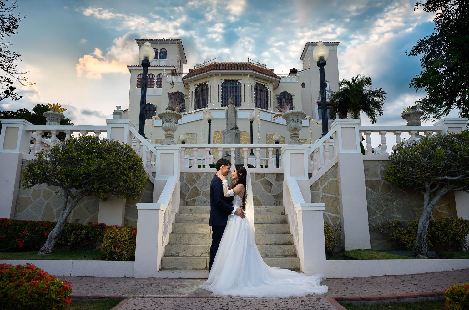 Bride and groom embrace on grand white staircase of elegant mansion venue under dramatic cloudy sky.