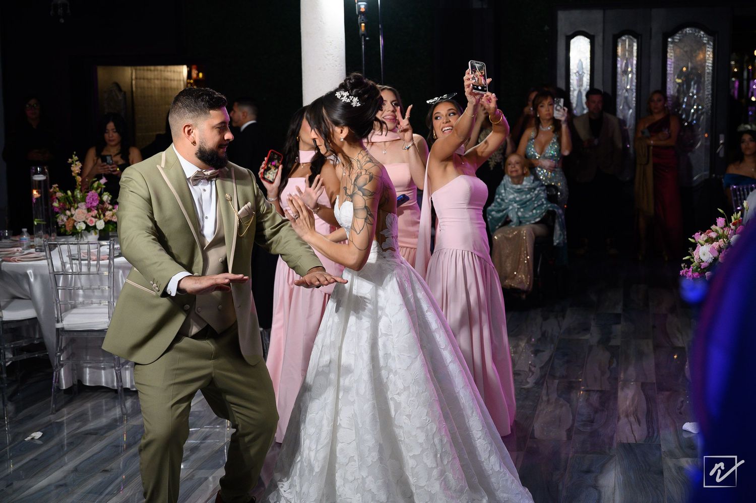 Bride in a white ball gown dances with a groom in an olive suit while bridesmaids in pink dresses cheer at an elegant wedding reception.