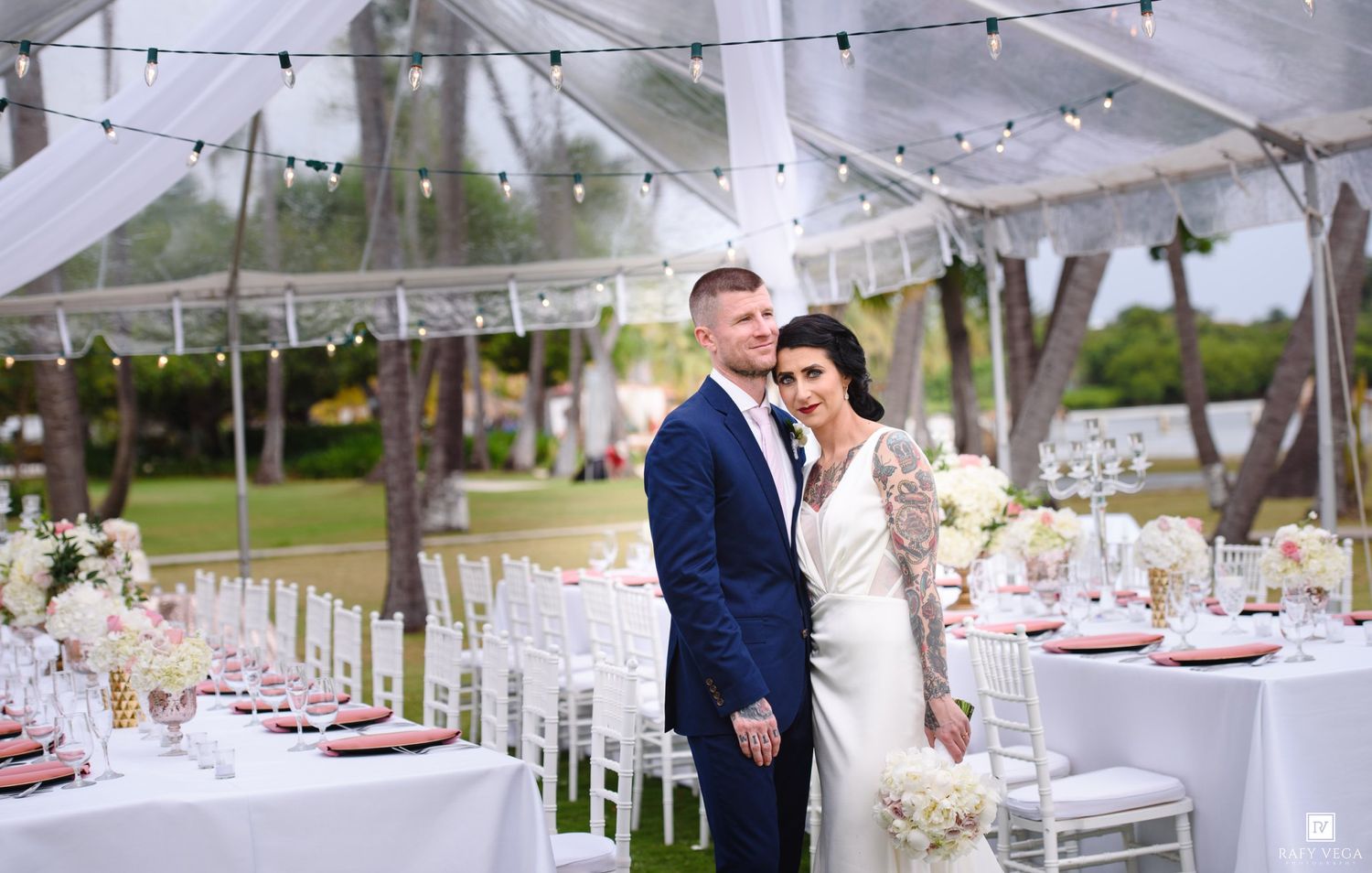 Bride and groom pose elegantly under a draped tent at a romantic outdoor wedding reception with white floral decor.