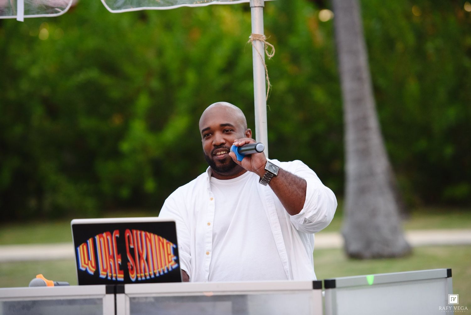 DJ in white outfit speaking into microphone at an outdoor tropical wedding event with palm trees in background.