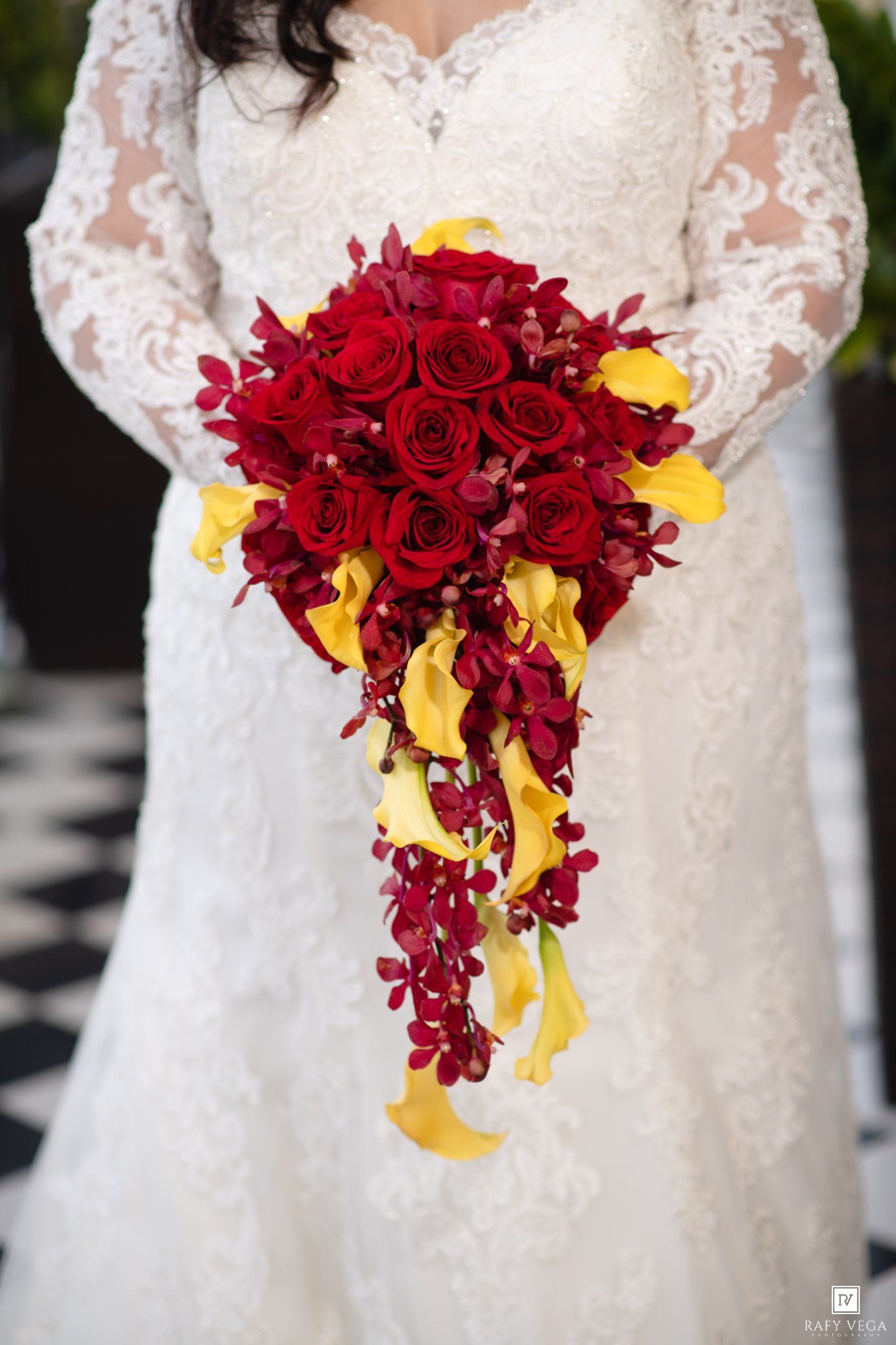 Bride in lace wedding gown holding cascading bouquet of red roses, burgundy orchids, and yellow calla lilies.