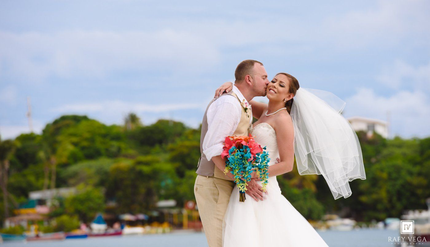 A bride and groom share a romantic kiss outdoors near a tropical waterfront on their wedding day.