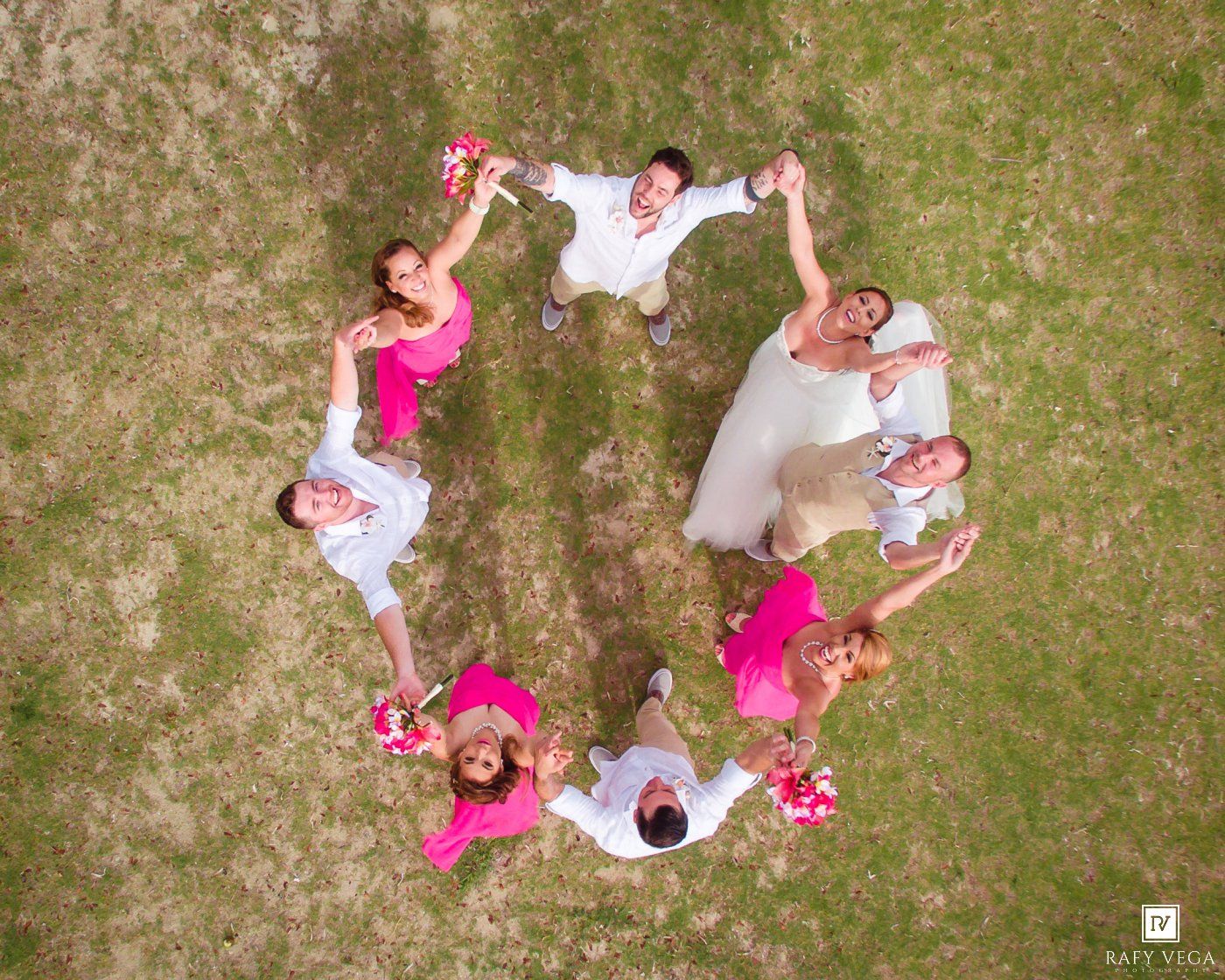 Aerial view of wedding party forming heart shape on grass with pink bridesmaid dresses.