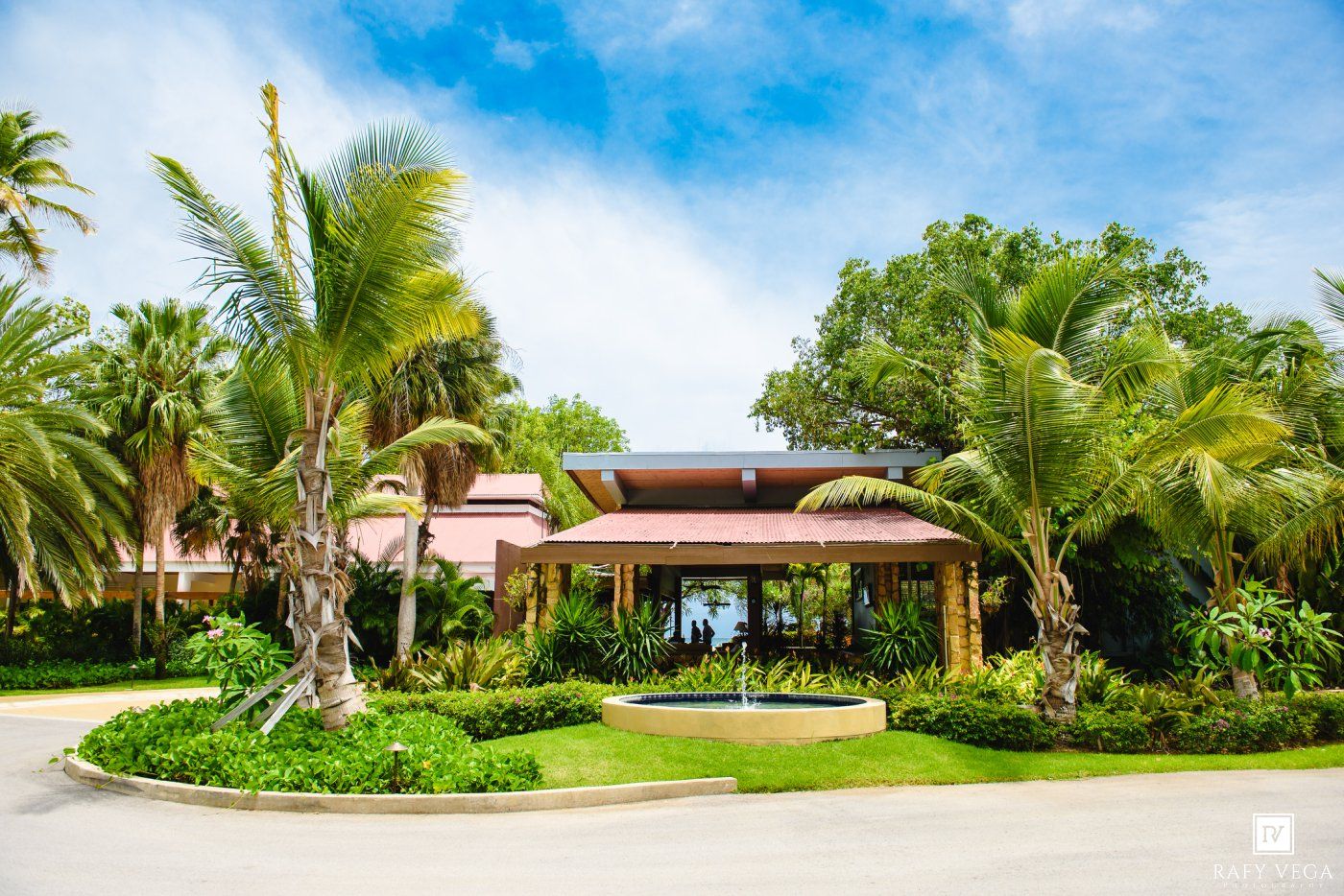 Tropical resort entrance with palm trees, lush gardens, fountain, and a pink building under a bright blue sky.