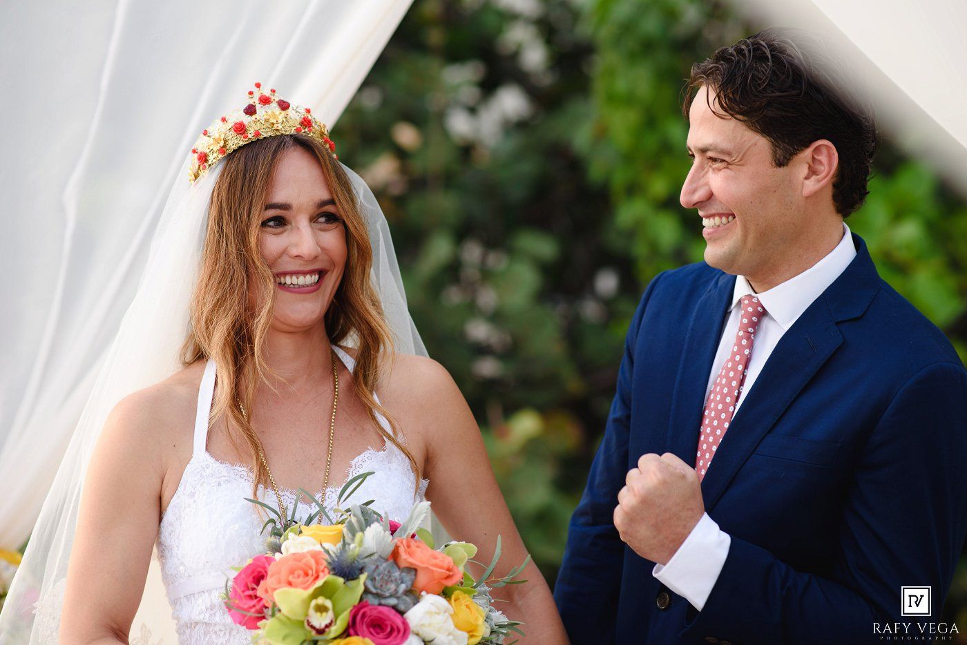 Smiling bride in white dress with floral crown holds colorful bouquet beside groom in navy suit at outdoor wedding.