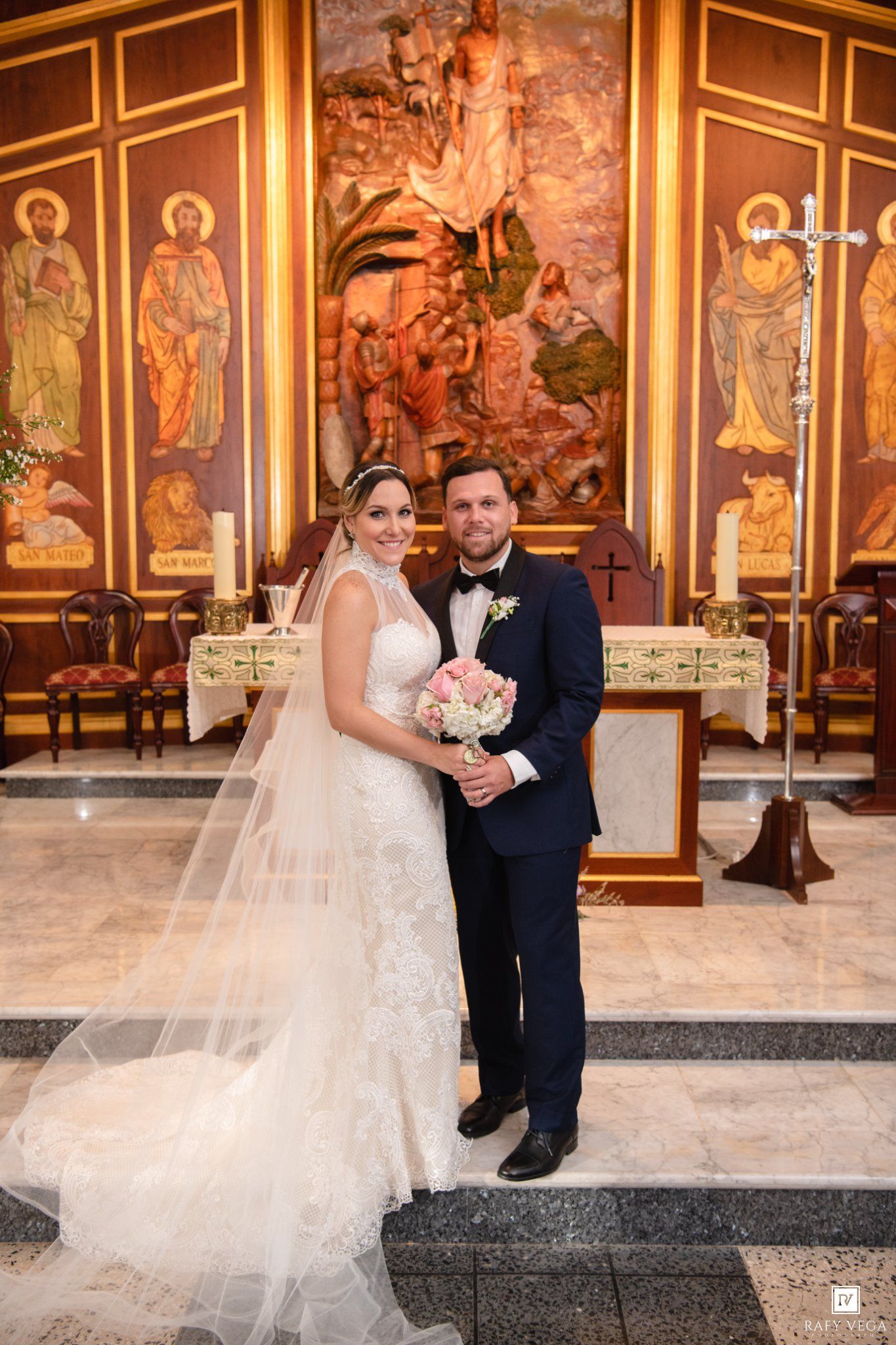 Bride and groom posing together at church altar, bride in lace gown with veil, groom in navy suit with bow tie.