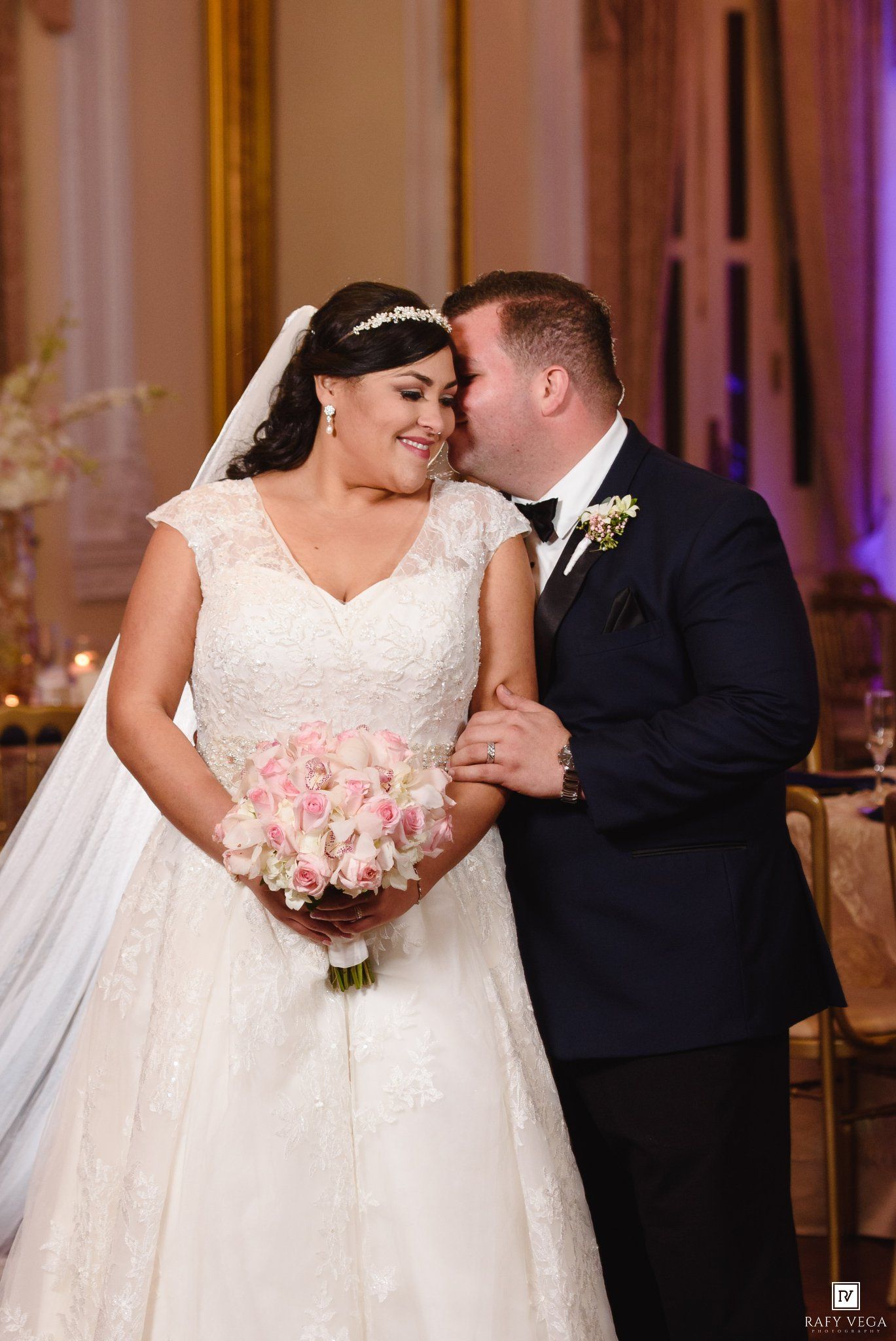 Bride in lace wedding gown holding pink rose bouquet shares tender moment with groom in navy tuxedo at elegant reception.