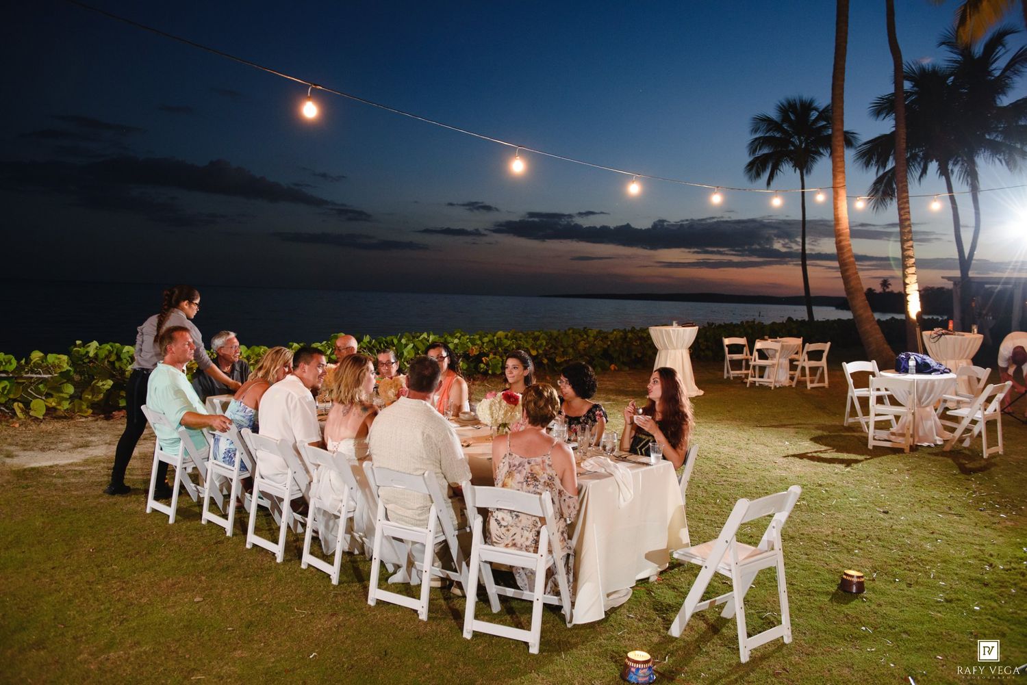 Outdoor wedding reception dinner on tropical beachfront lawn at sunset with string lights and white chairs.