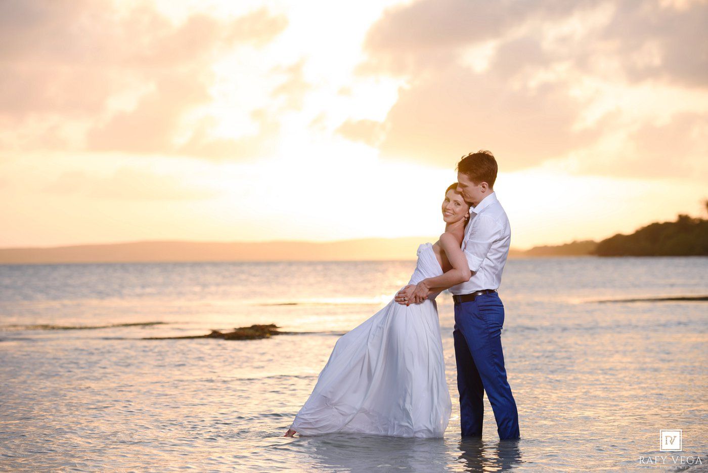 Couple embracing on a tropical beach at sunset, bride in white gown and groom in blue pants, ocean waves at their feet.