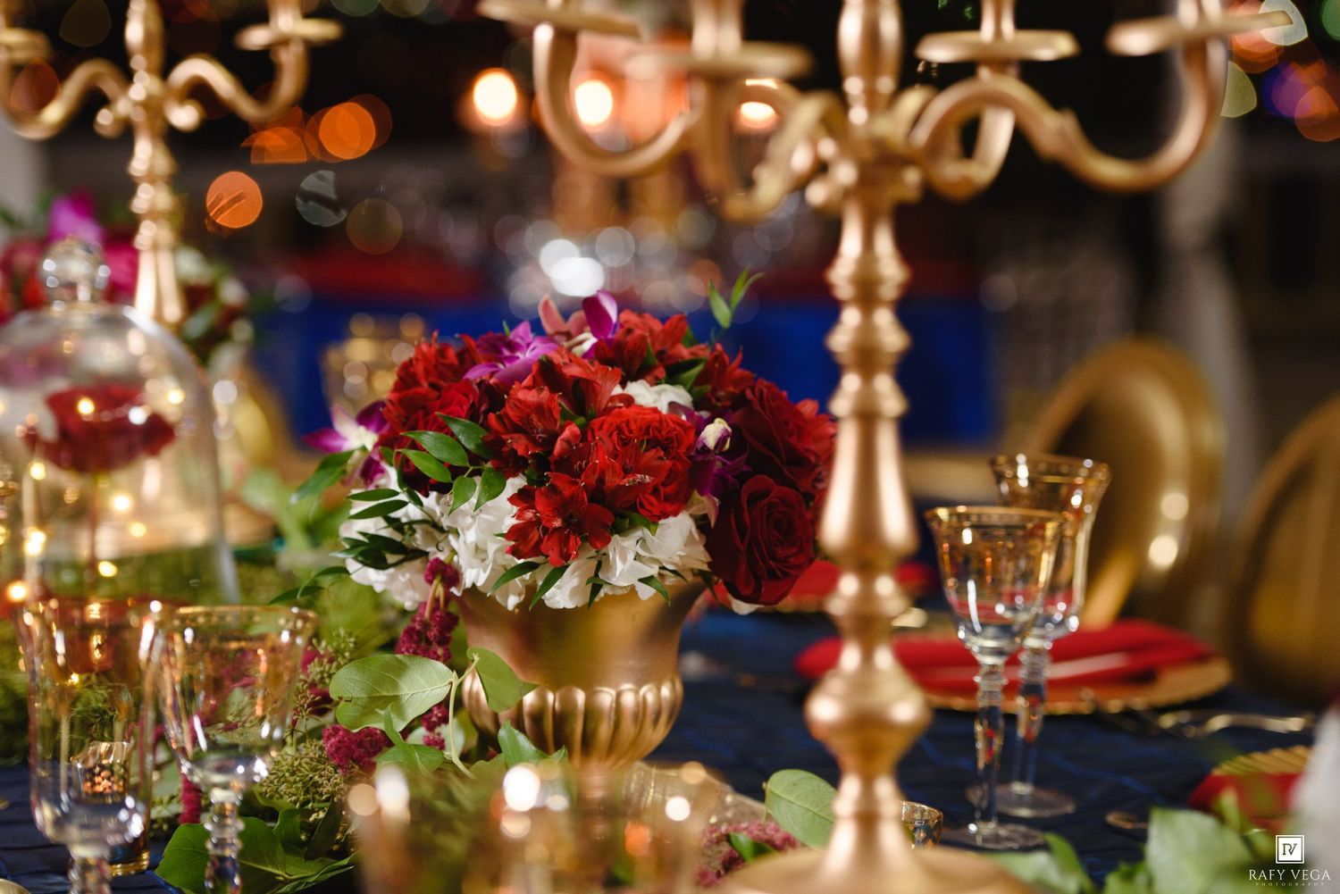 Elegant wedding table centerpiece with red roses in gold vase, crystal glasses, and gold candelabras on navy blue tablecloth.