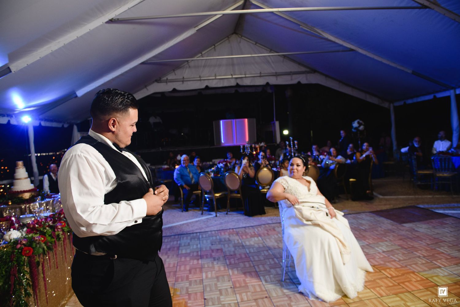 Bride and groom share first dance under white tent at elegant outdoor wedding reception with guests watching.