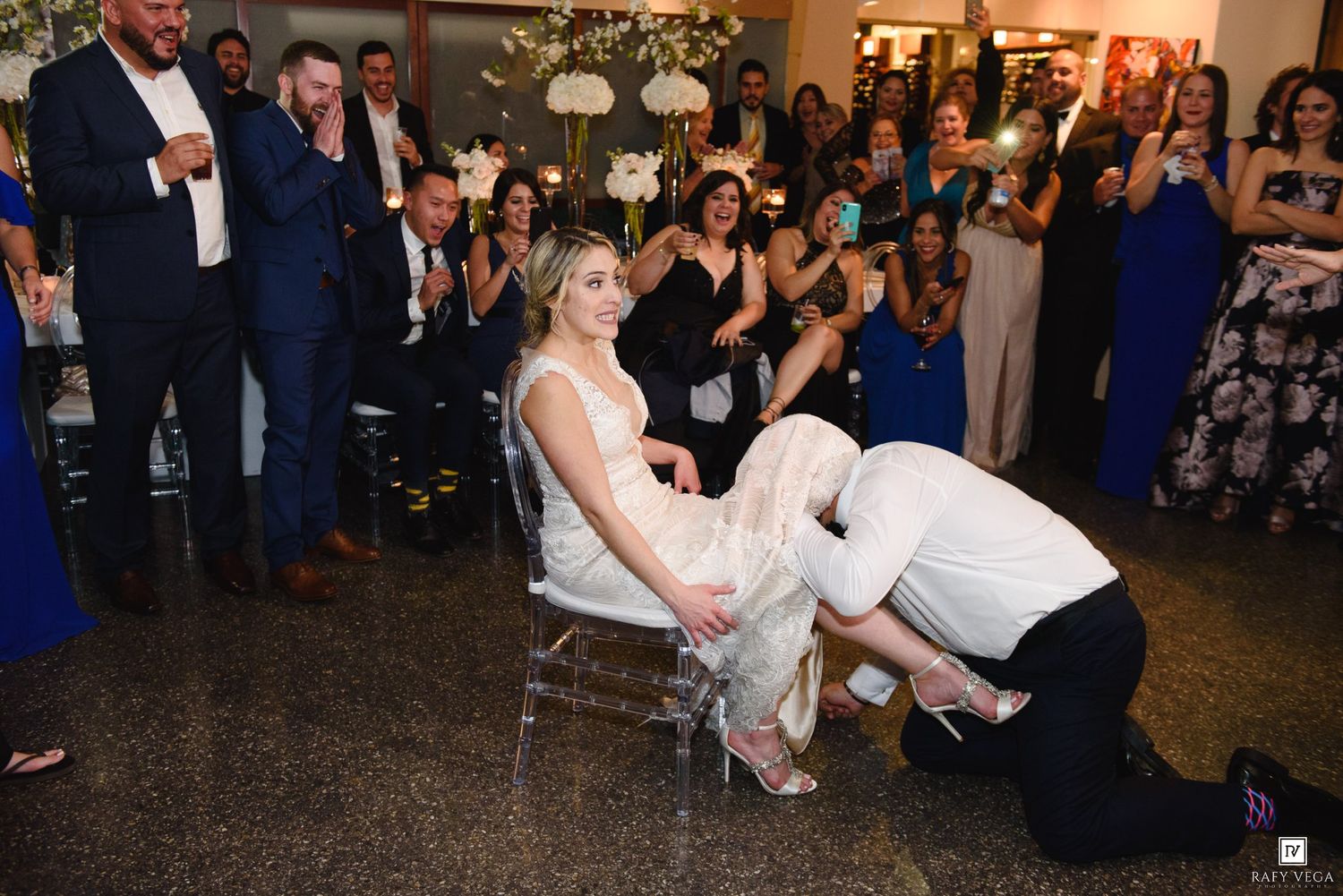 Groom removes garter from bride seated in chair at wedding reception while guests cheer and watch.