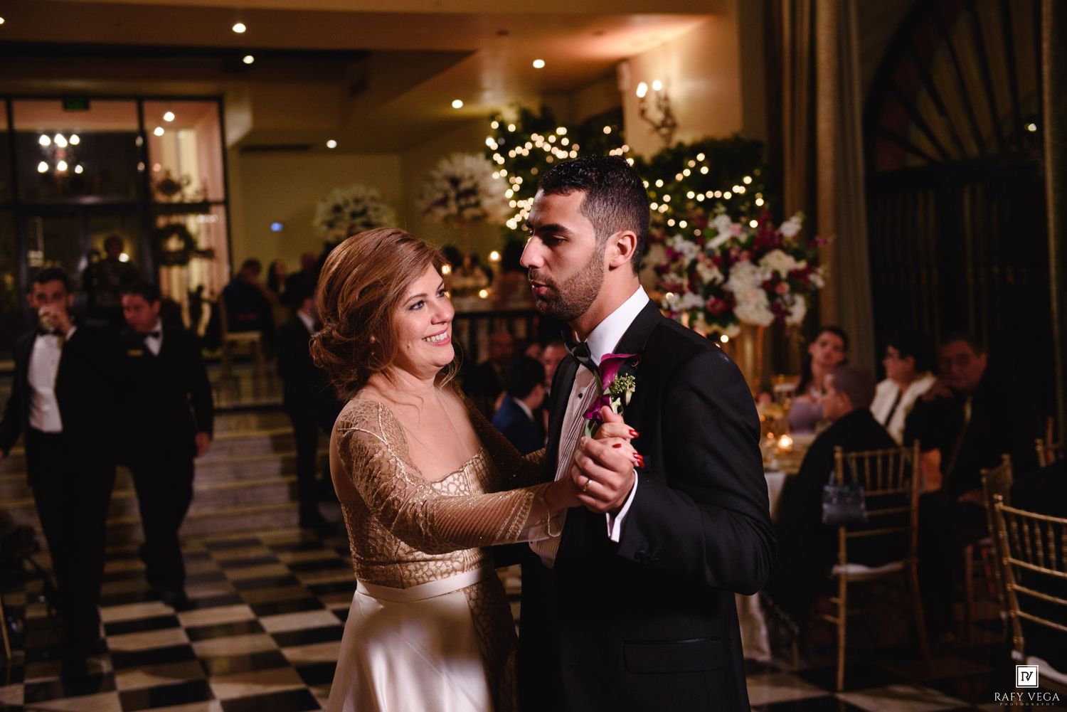 Groom shares a mother-son dance with his mom at an elegant wedding reception with warm lighting.