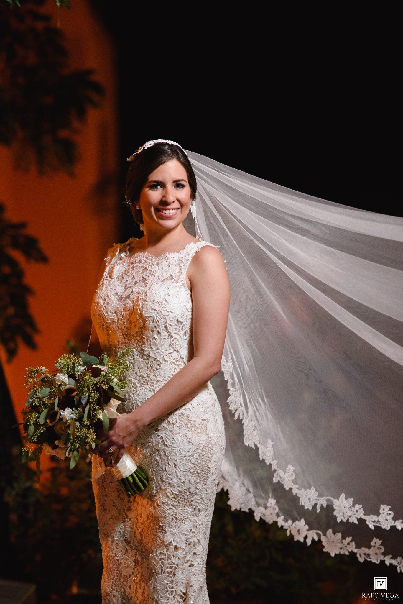 Bride in lace mermaid wedding dress holding bouquet with flowing veil, smiling elegantly at outdoor night ceremony.
