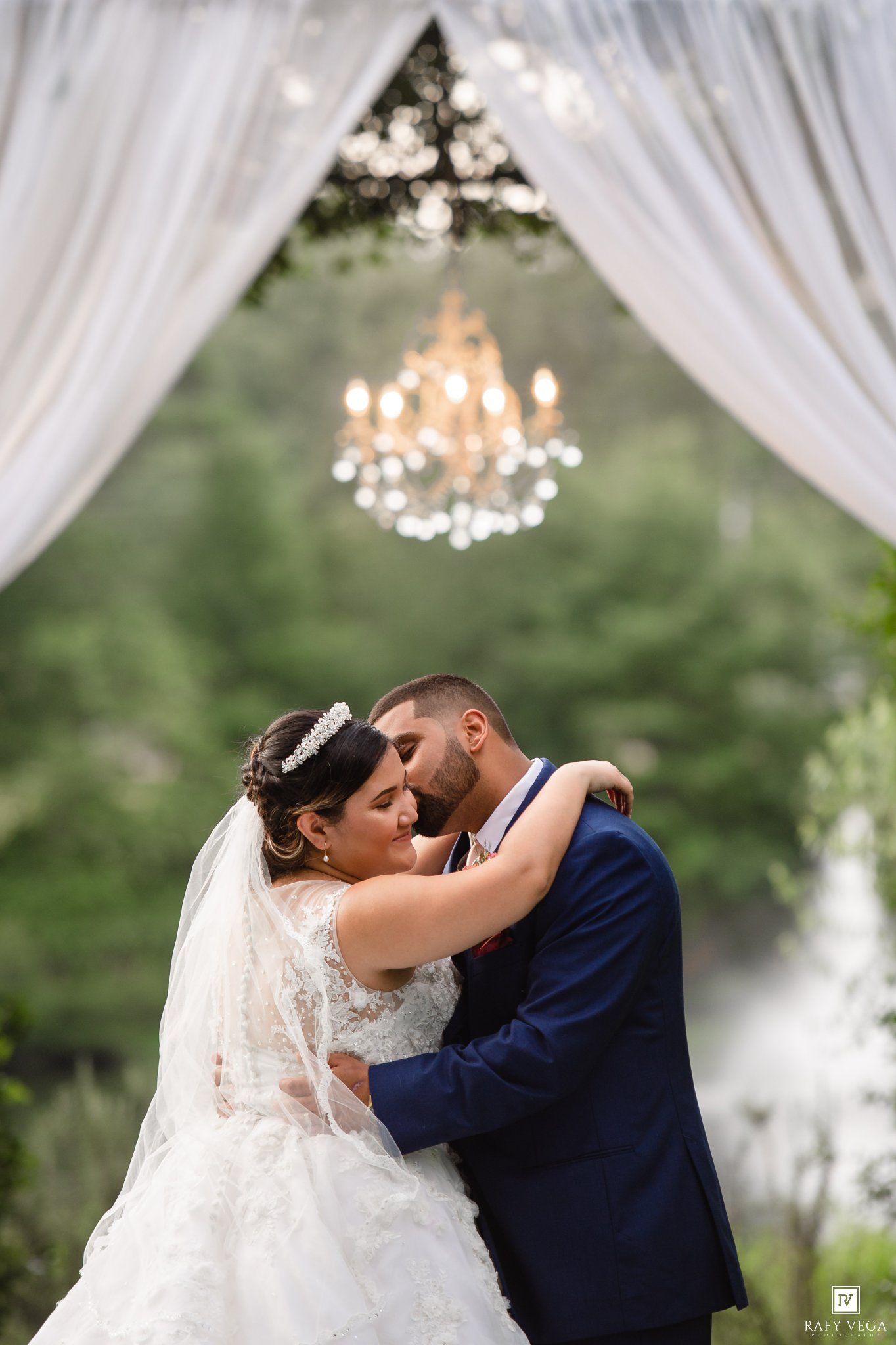 Bride and groom share romantic embrace under chandelier and white draped arch at outdoor wedding ceremony.