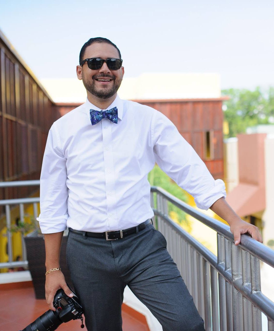 Man in white shirt and blue bow tie holding camera, smiling on sunny outdoor balcony with sunglasses.