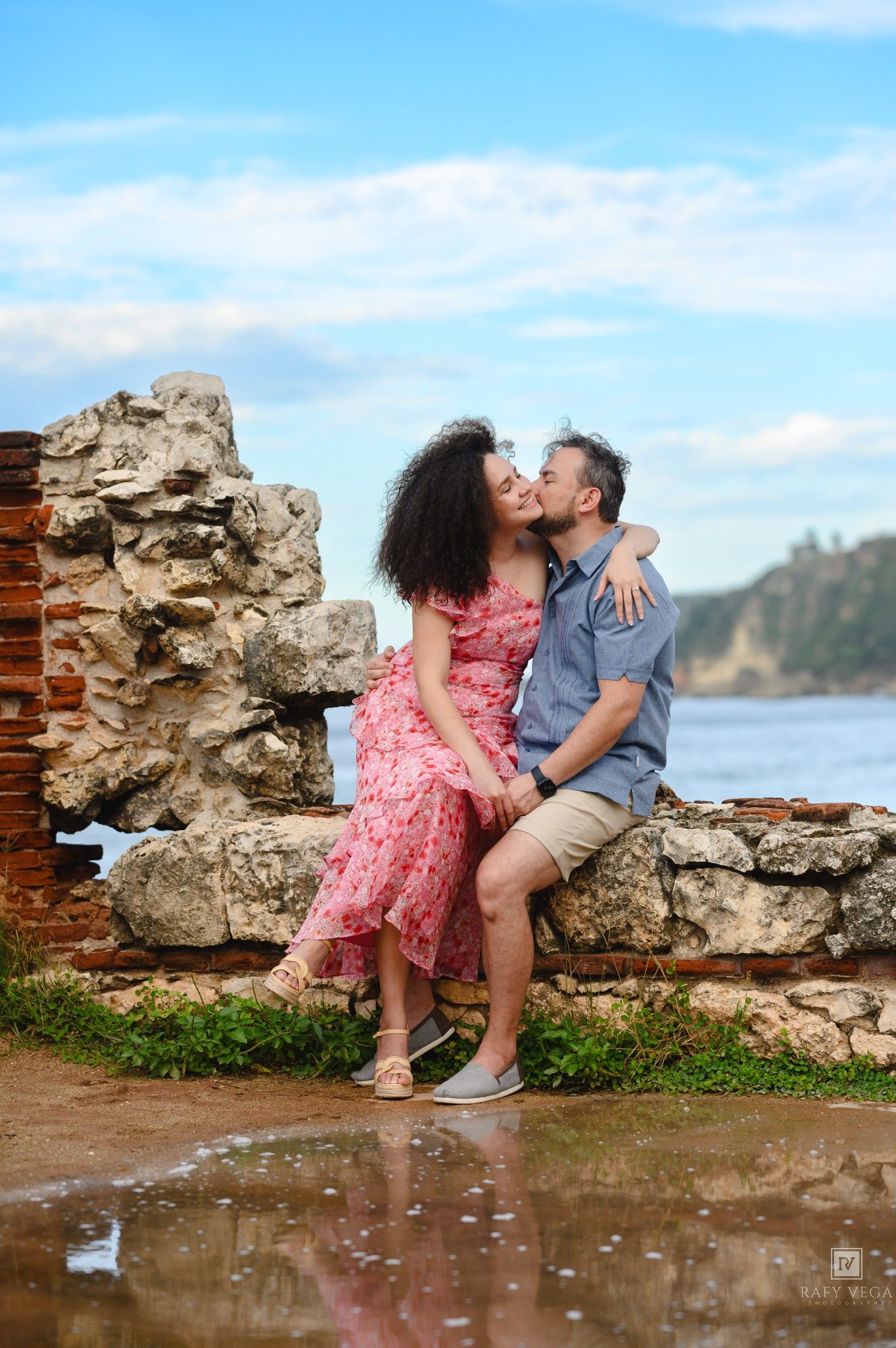 Romantic couple sharing a kiss on ancient stone ruins by the sea, woman in pink floral dress, man in blue shirt.
