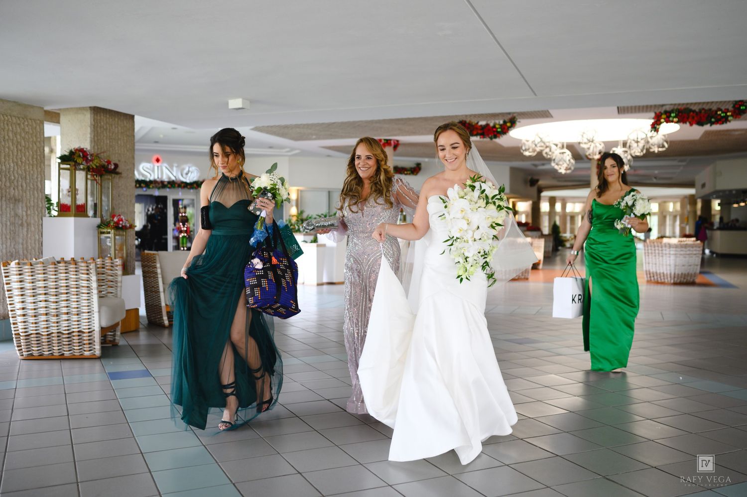 Bride in white gown walks with bridesmaids in green and silver dresses through elegant venue hall.