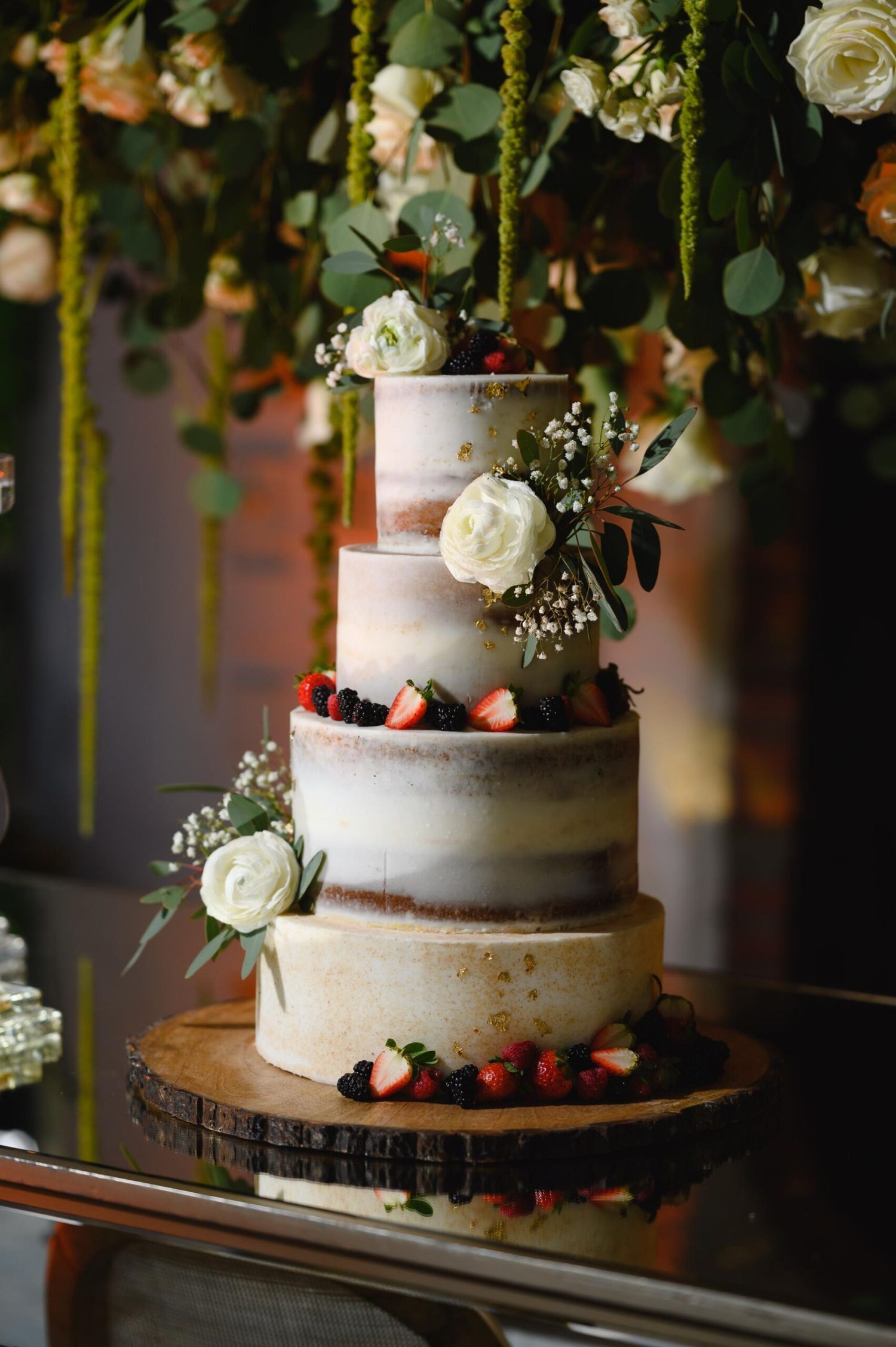 Three-tier semi-naked wedding cake with fresh berries, white roses, and greenery on a rustic wood slice stand.