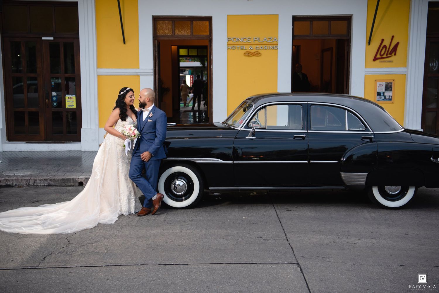 Wedding couple posing beside a classic black vintage car on a colorful street in front of a yellow building.