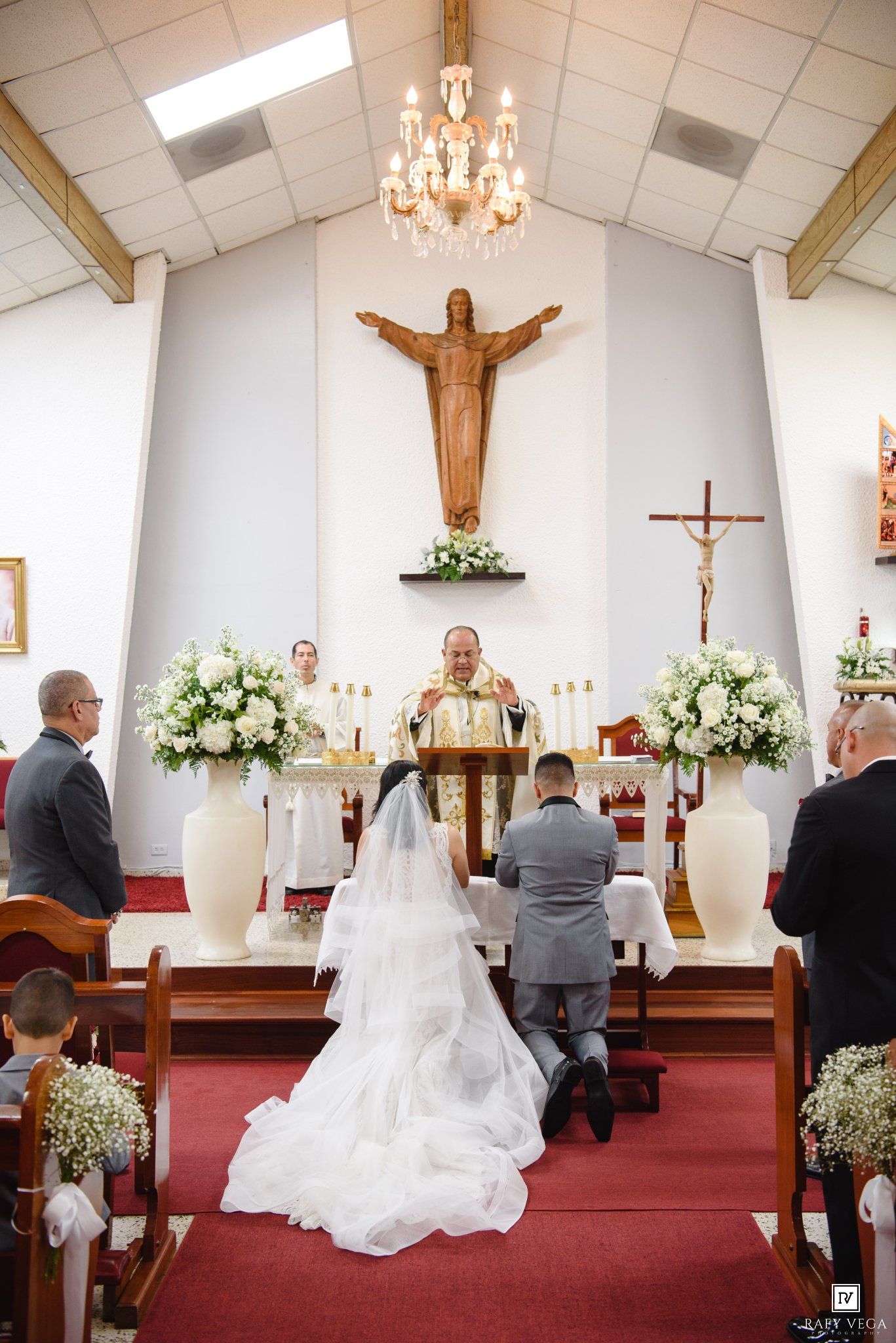 Bride and groom kneel at church altar during Catholic wedding ceremony with priest officiating.