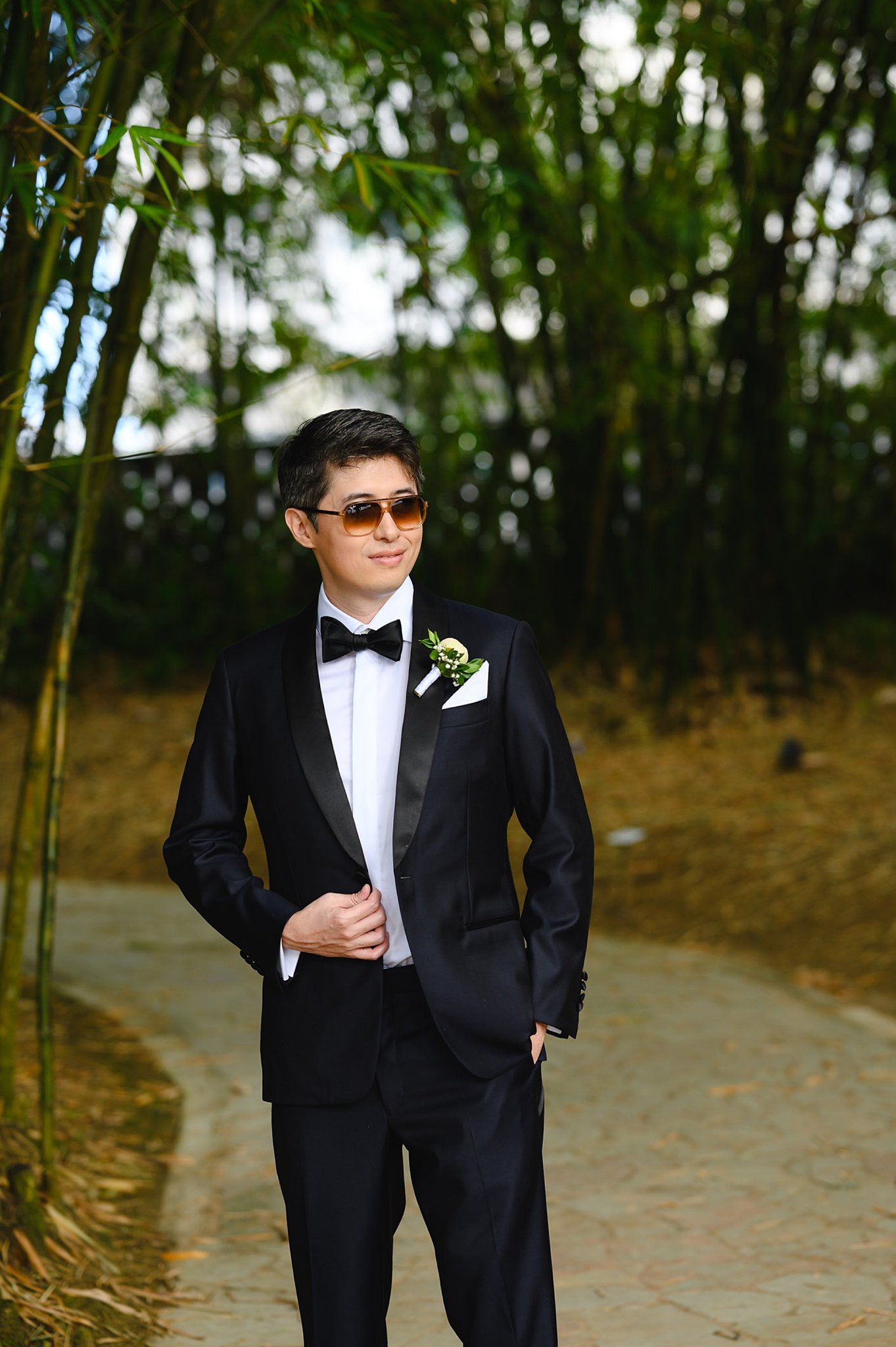 Groom in black tuxedo with bow tie and boutonniere wearing sunglasses, posing outdoors among bamboo trees.