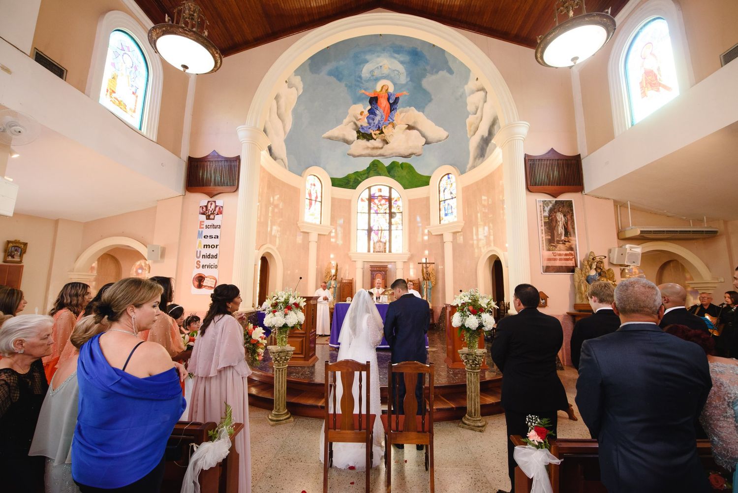 Wedding ceremony inside ornate church with mural, guests standing as couple exchanges vows at altar.