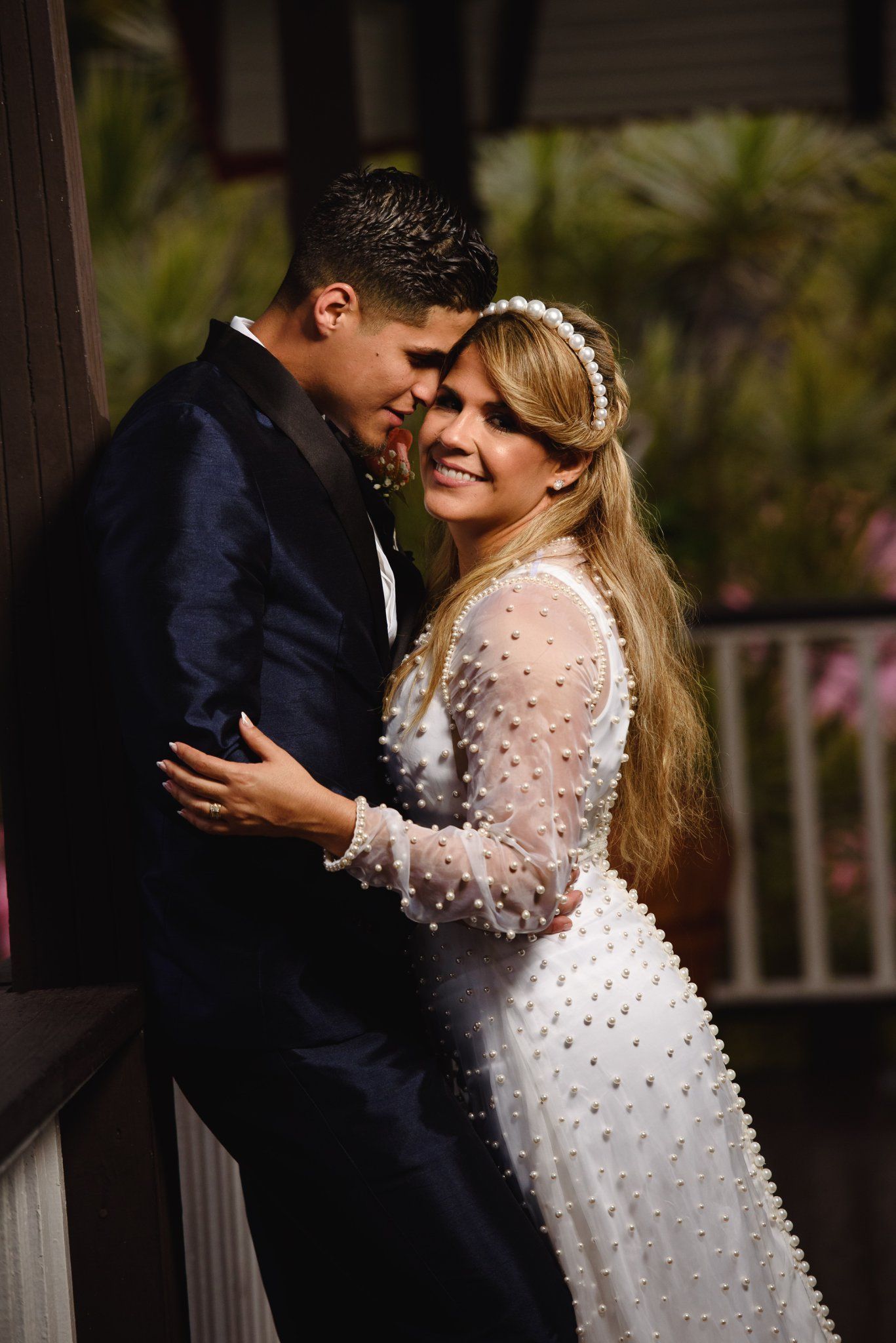 Bride in white pearl gown and groom in navy suit share romantic embrace, smiling together at outdoor wedding.