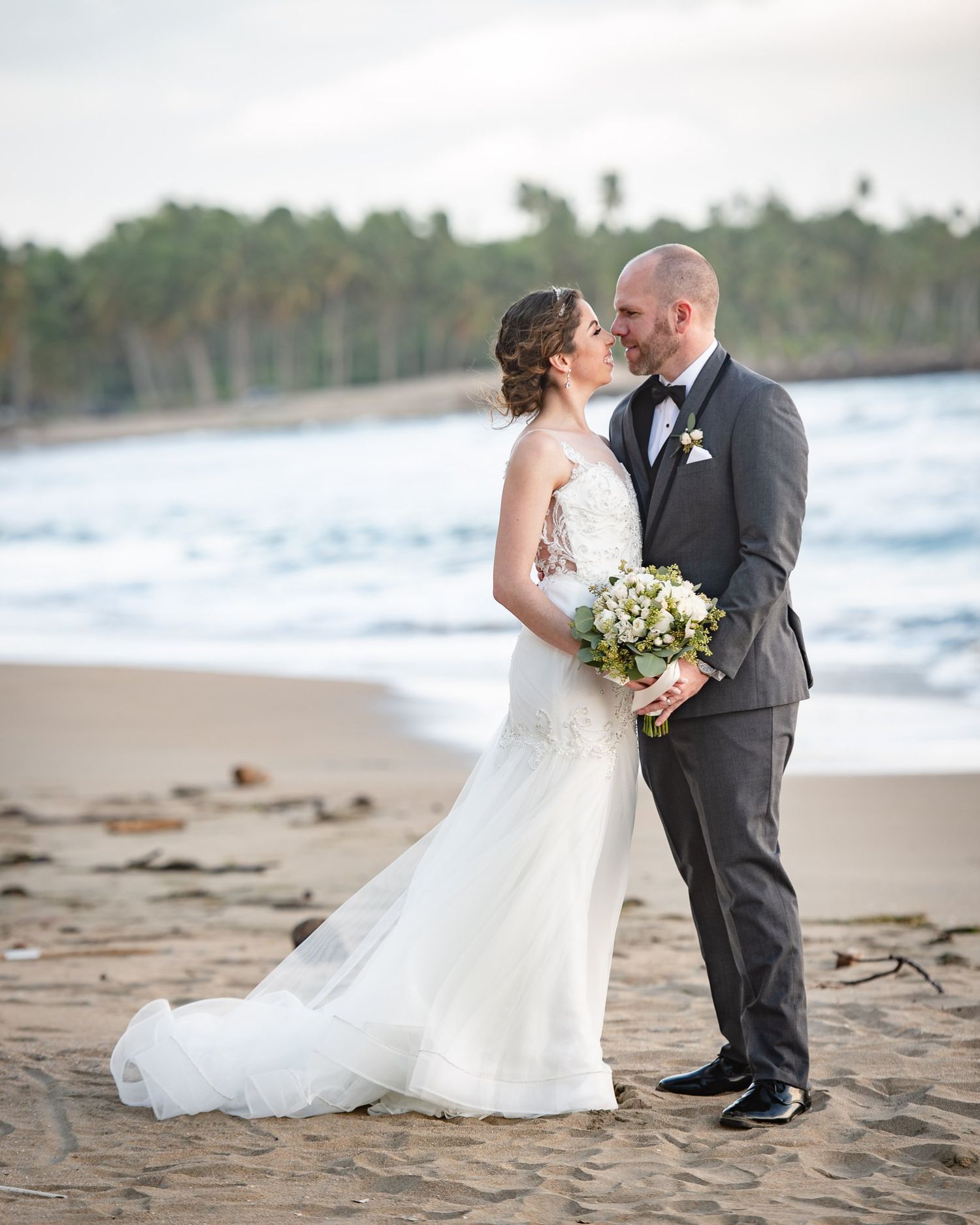 Bride and groom sharing a romantic kiss on a sandy beach, bride holding a white bouquet in a lace wedding gown.