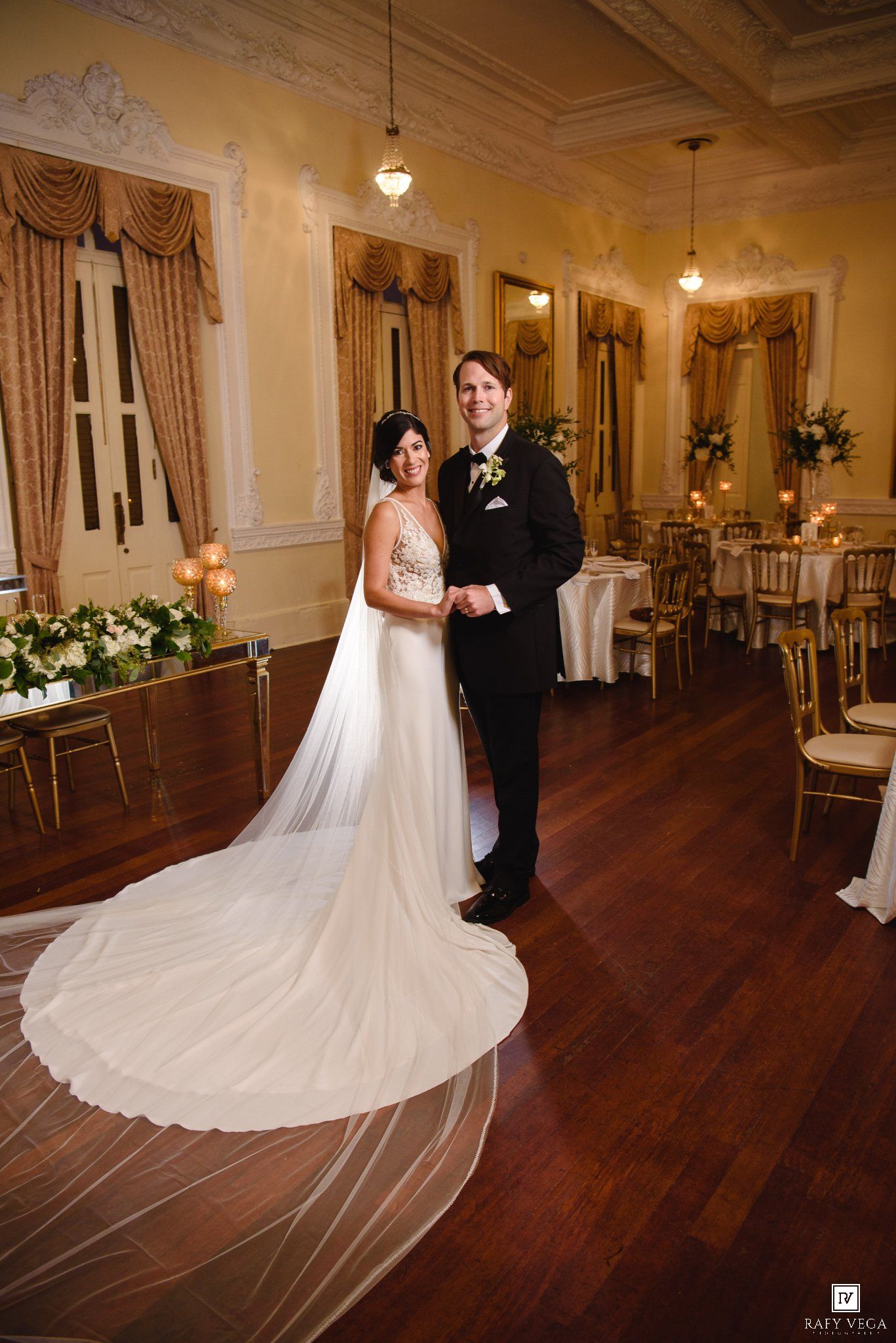 Bride in flowing veil and groom in tuxedo pose in ornate ballroom reception venue.