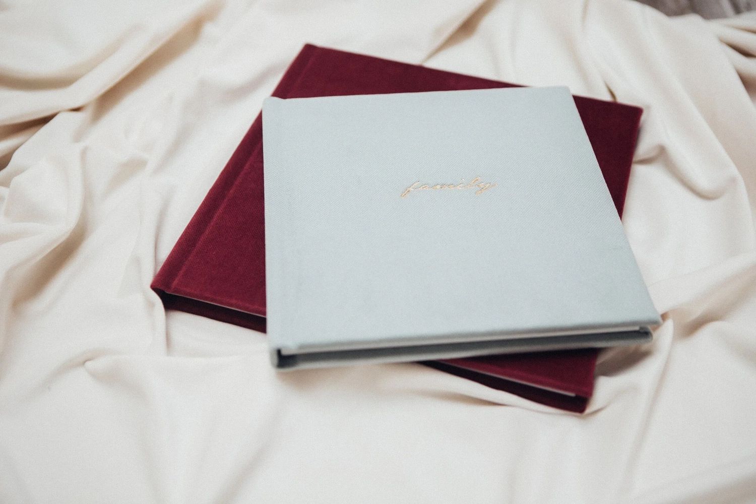 Two elegant velvet photo albums in light blue and burgundy stacked on white silk fabric.