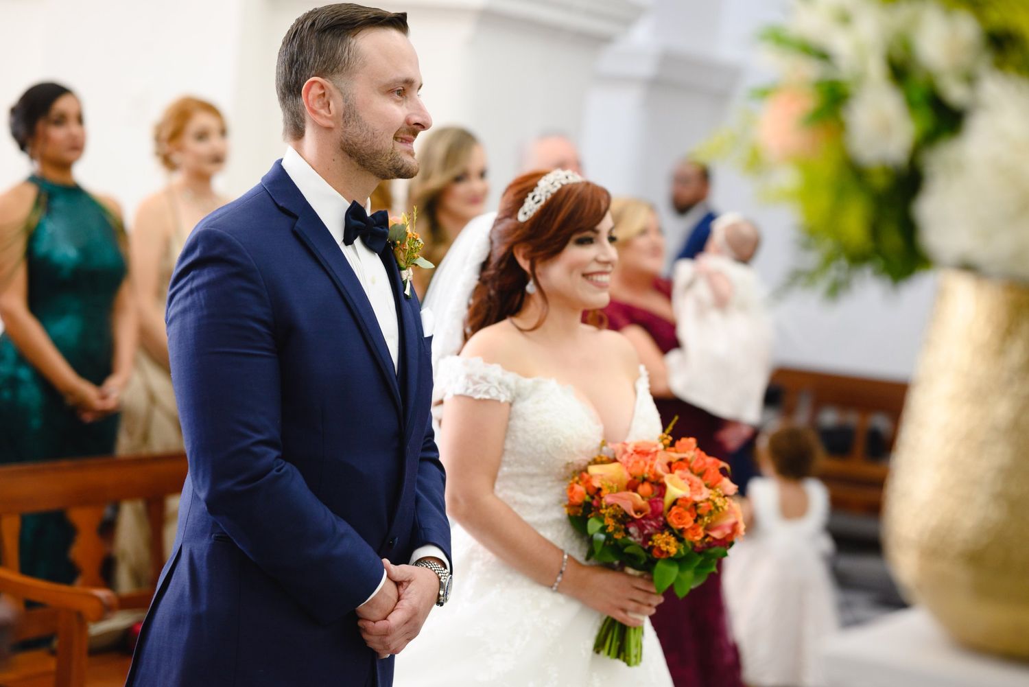 Groom in navy suit stands with bride in white lace gown holding orange bouquet at church wedding.