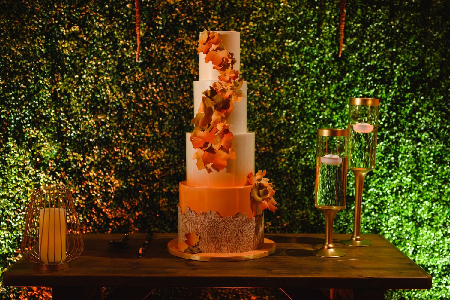 Elegant four-tier autumn wedding cake with orange floral decor on a wooden table against a lush green hedge backdrop.