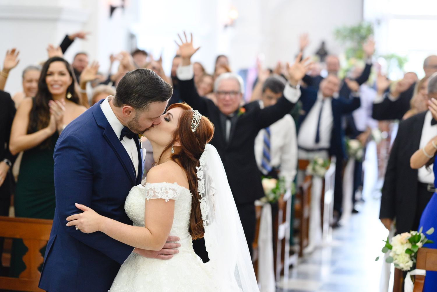 Bride and groom share first kiss at church altar as joyful wedding guests celebrate with raised arms behind them.