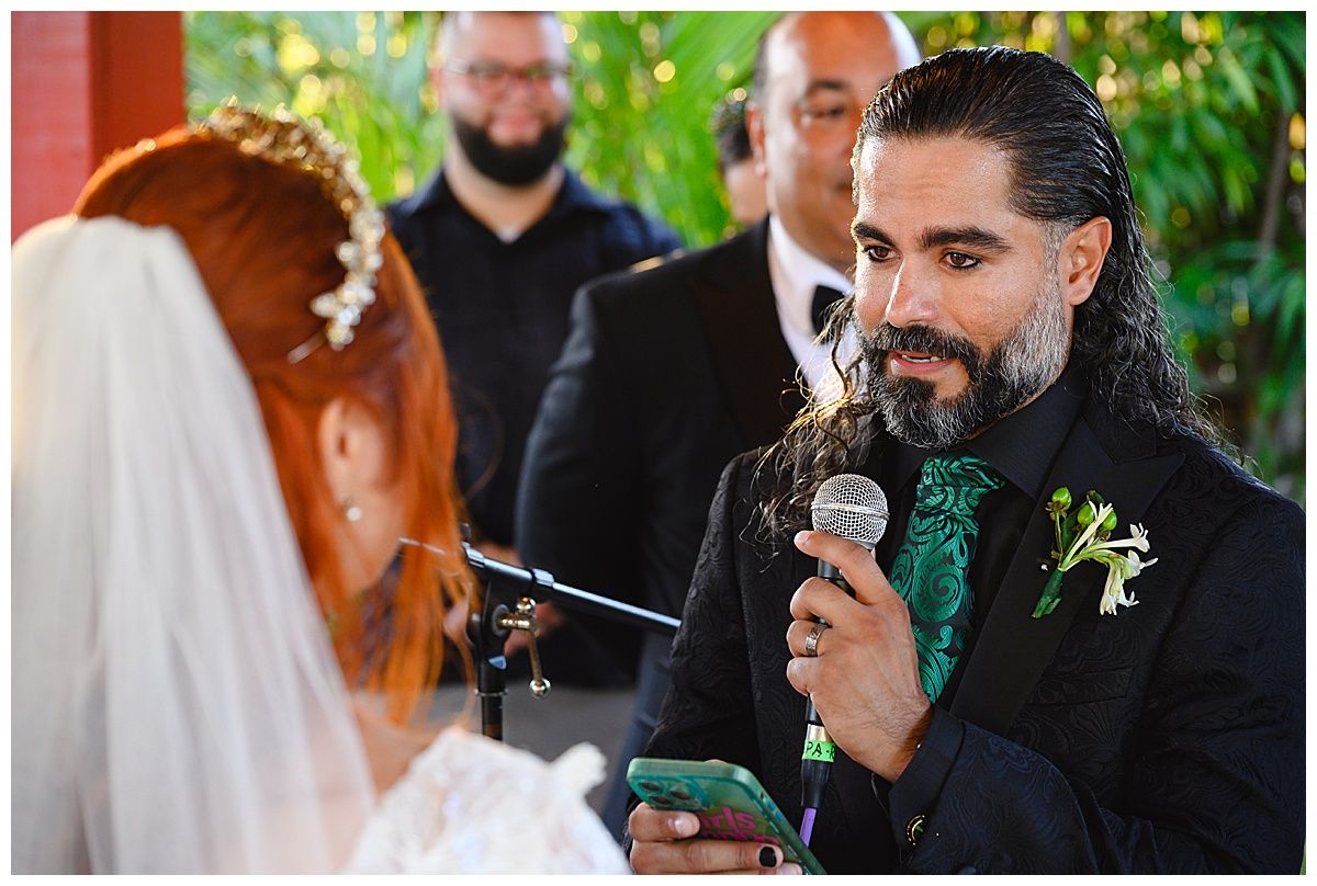 Groom in black suit with green tie reads vows from phone to red-haired bride outdoors.
