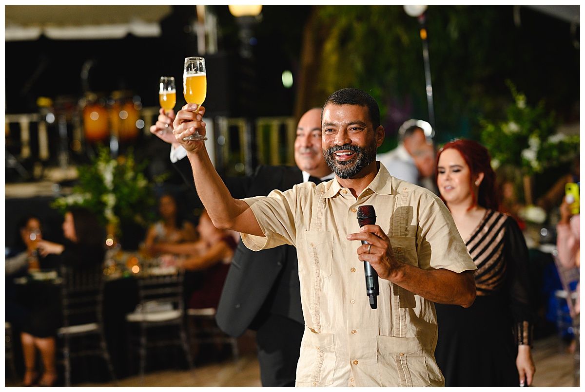 Man in beige suit raises champagne glass while giving a toast at an elegant outdoor wedding reception.