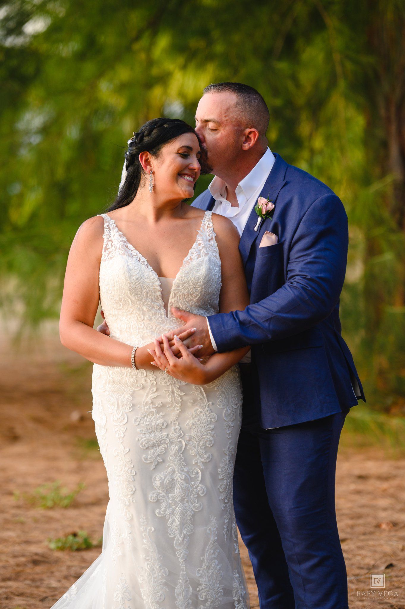 Bride in lace gown and groom in blue suit share a tender kiss outdoors with green trees in background.