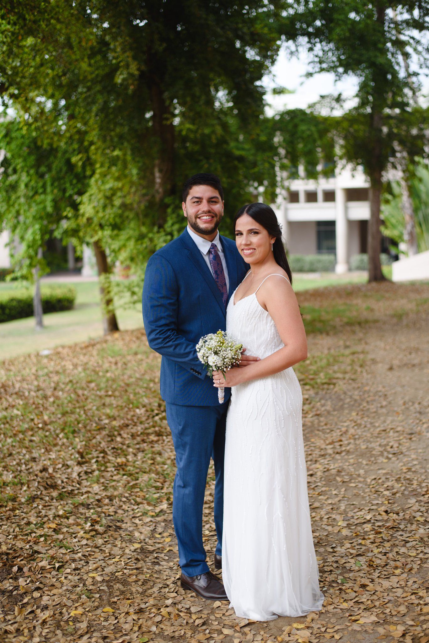 Smiling bride and groom pose outdoors in a park, she holds a small bouquet in a white gown, he wears a navy suit.