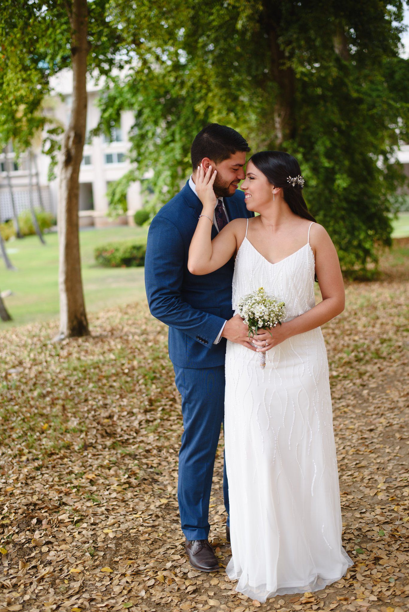 Bride in white gown holding bouquet and groom in blue suit share romantic moment outdoors among autumn leaves.