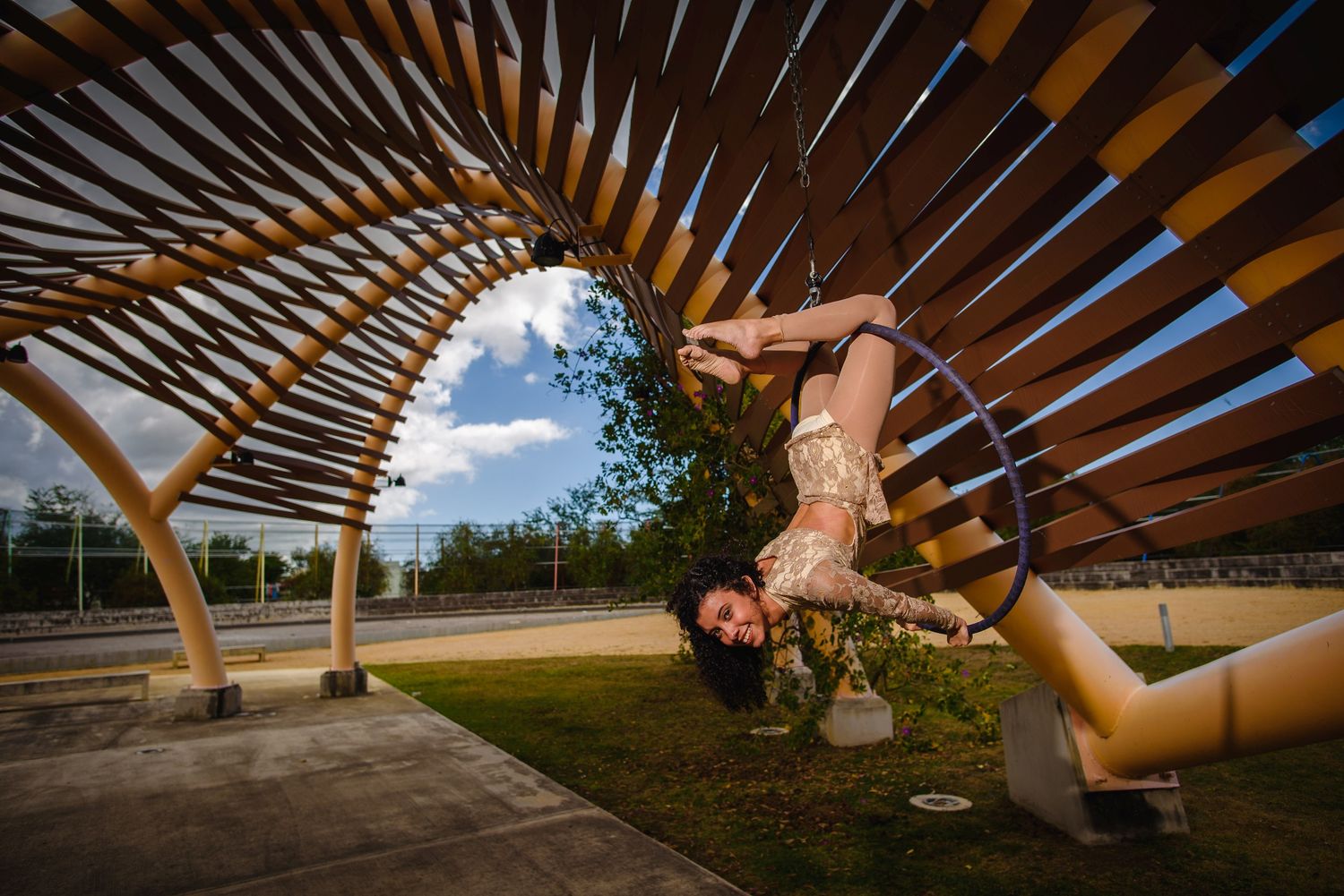 A woman performs an acrobatic pose with a hoop under a wooden arch structure in an outdoor park setting.