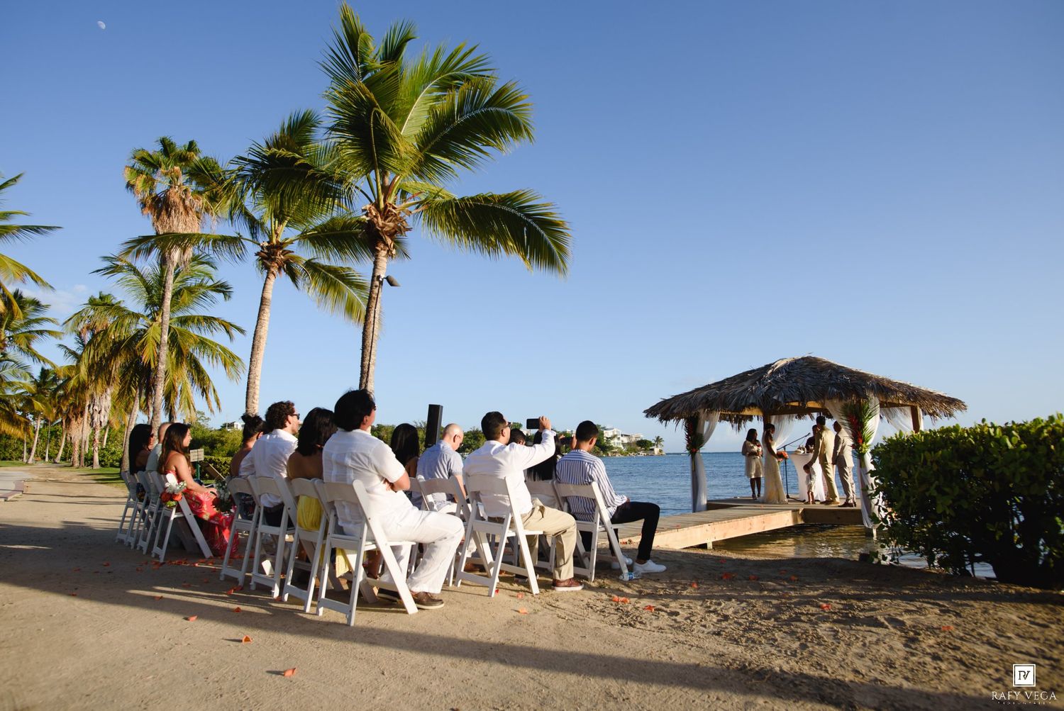Outdoor beach wedding ceremony with guests seated on white chairs under palm trees near a gazebo by the ocean.