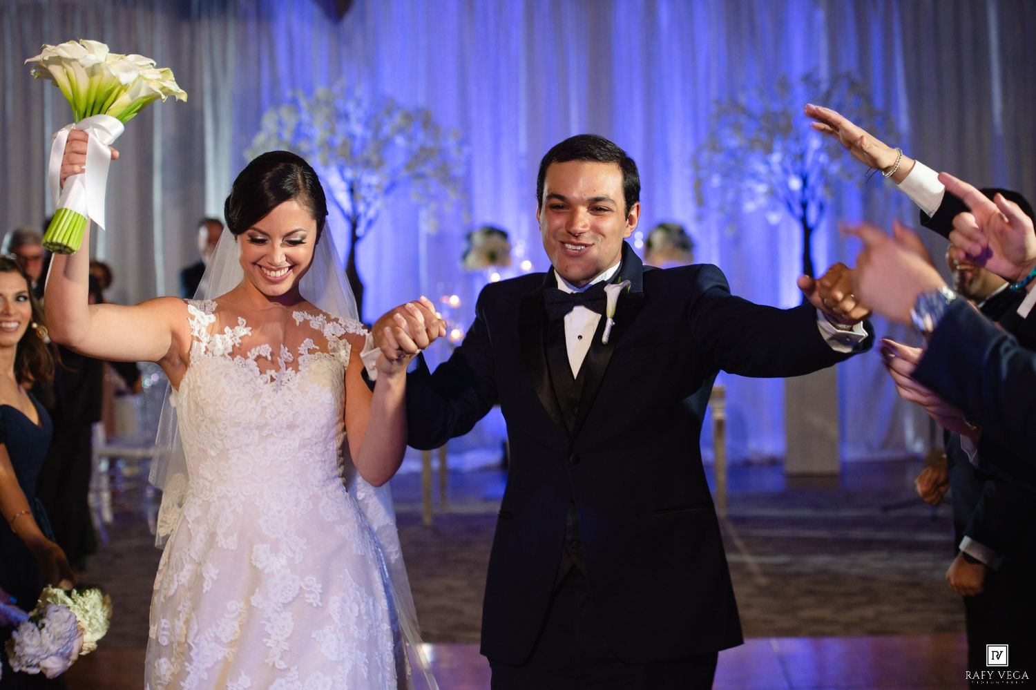 Joyful newlyweds celebrate their wedding ceremony exit, bride in lace gown holding calla lilies, groom in black tuxedo.