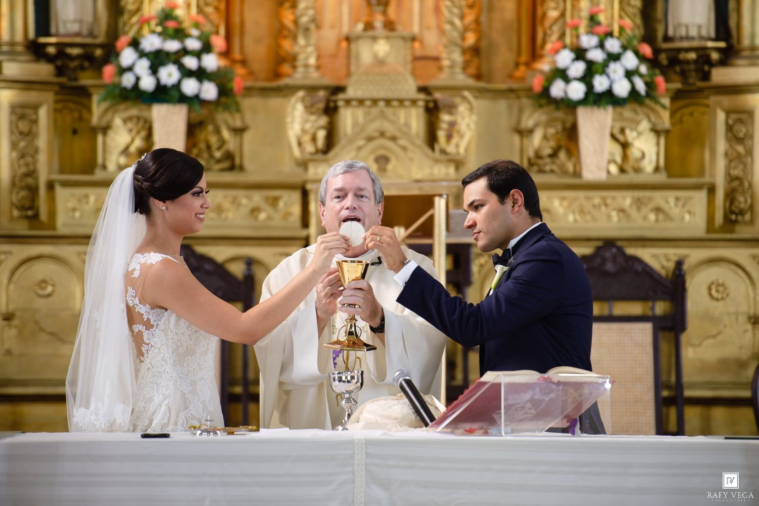 Bride and groom holding a chalice with a priest during a Catholic wedding ceremony at an ornate church altar.