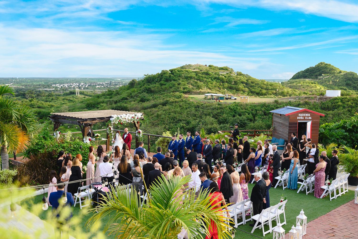 Outdoor wedding ceremony on a hilltop venue with lush green hills, blue sky, white chairs, and tropical landscape backdrop.