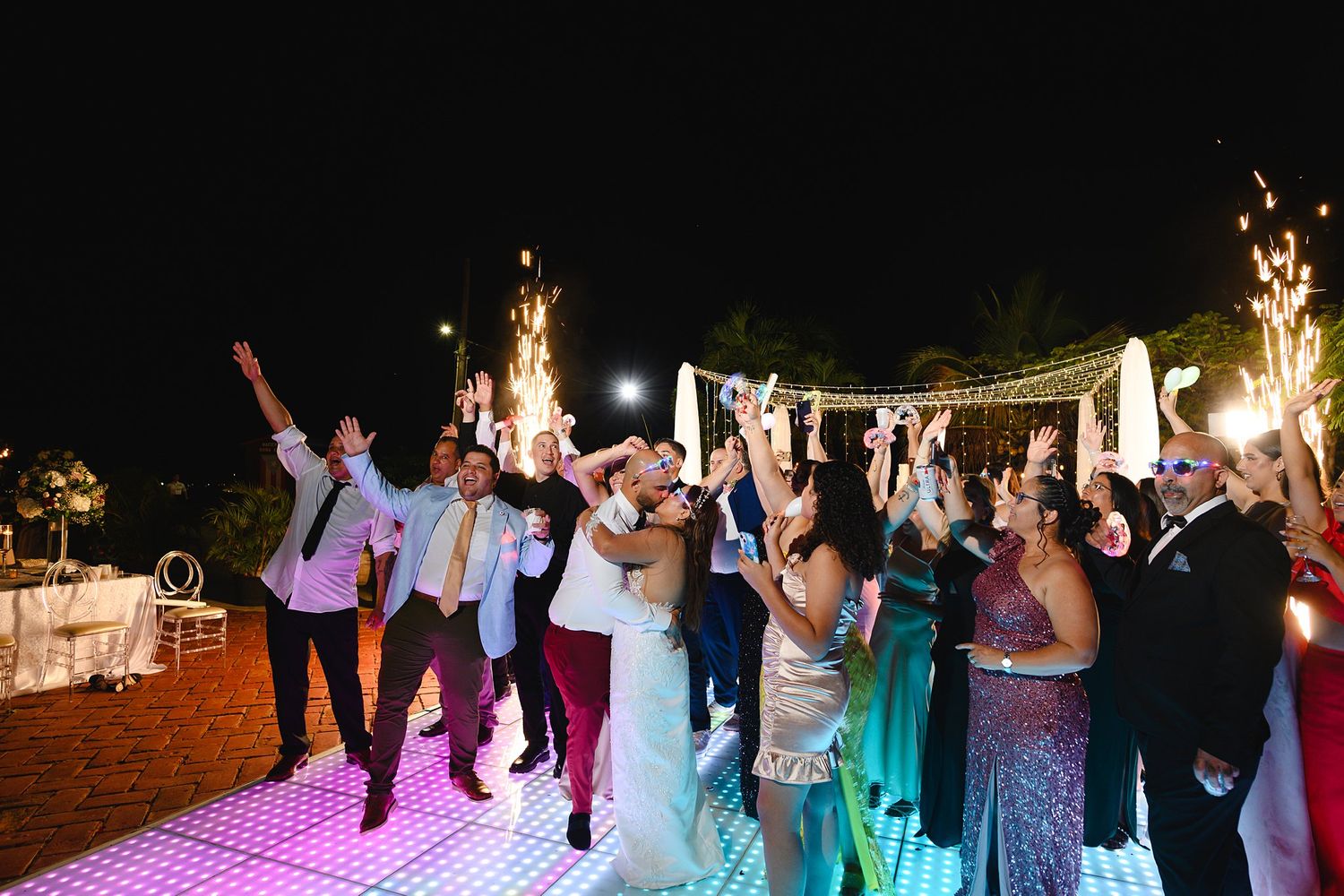 Wedding guests celebrate on a colorful dance floor at night with sparklers lighting up the festive outdoor reception.
