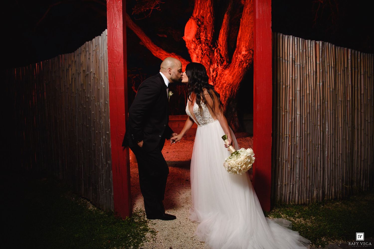 Bride and groom share a romantic kiss in front of a red-lit tree and wooden fence at night.