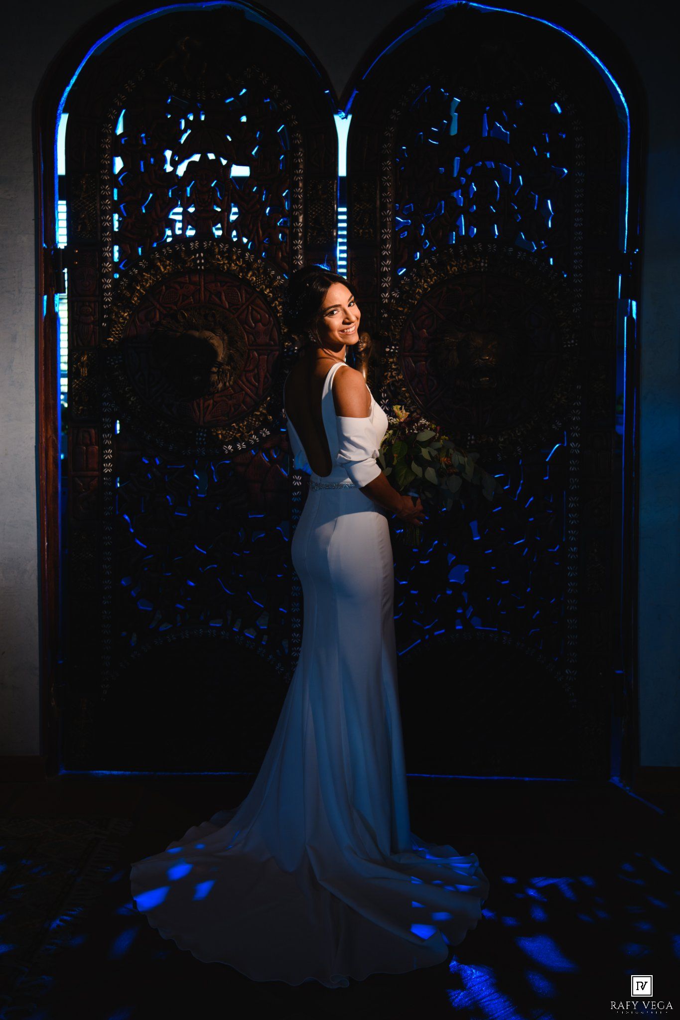 Bride in white mermaid gown holding bouquet poses before ornate dark doors with blue lighting.